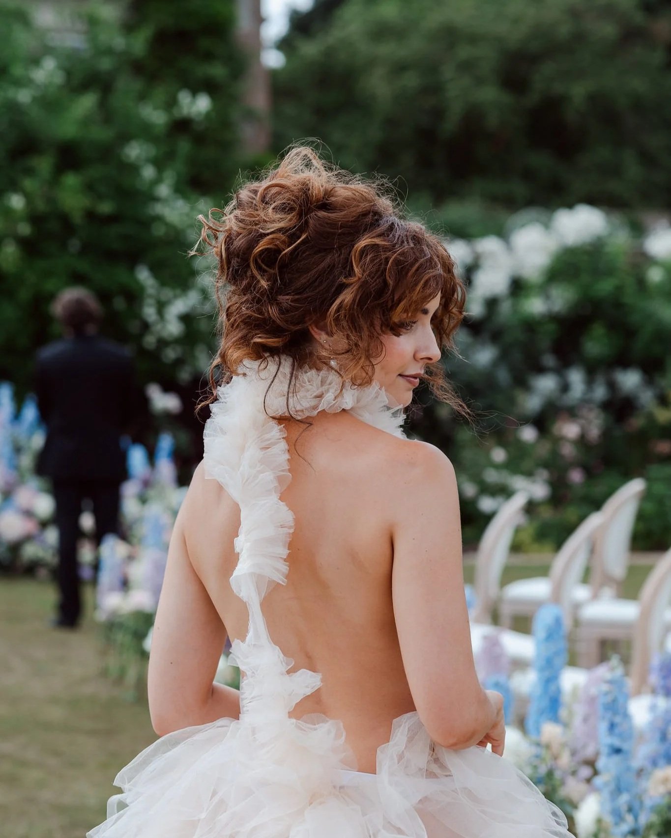 A woman with curly red hair wearing a white ruffled dress, outdoors at a garden or park, with chairs and flowers in the background.
