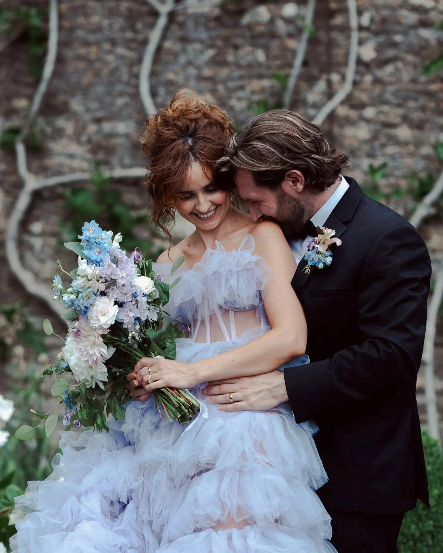 Bride in a ruffled white gown holding a bouquet of pastel-colored flowers, smiling with groom in a black suit. They are close, with the groom leaning in and smiling. Stone and vine background.