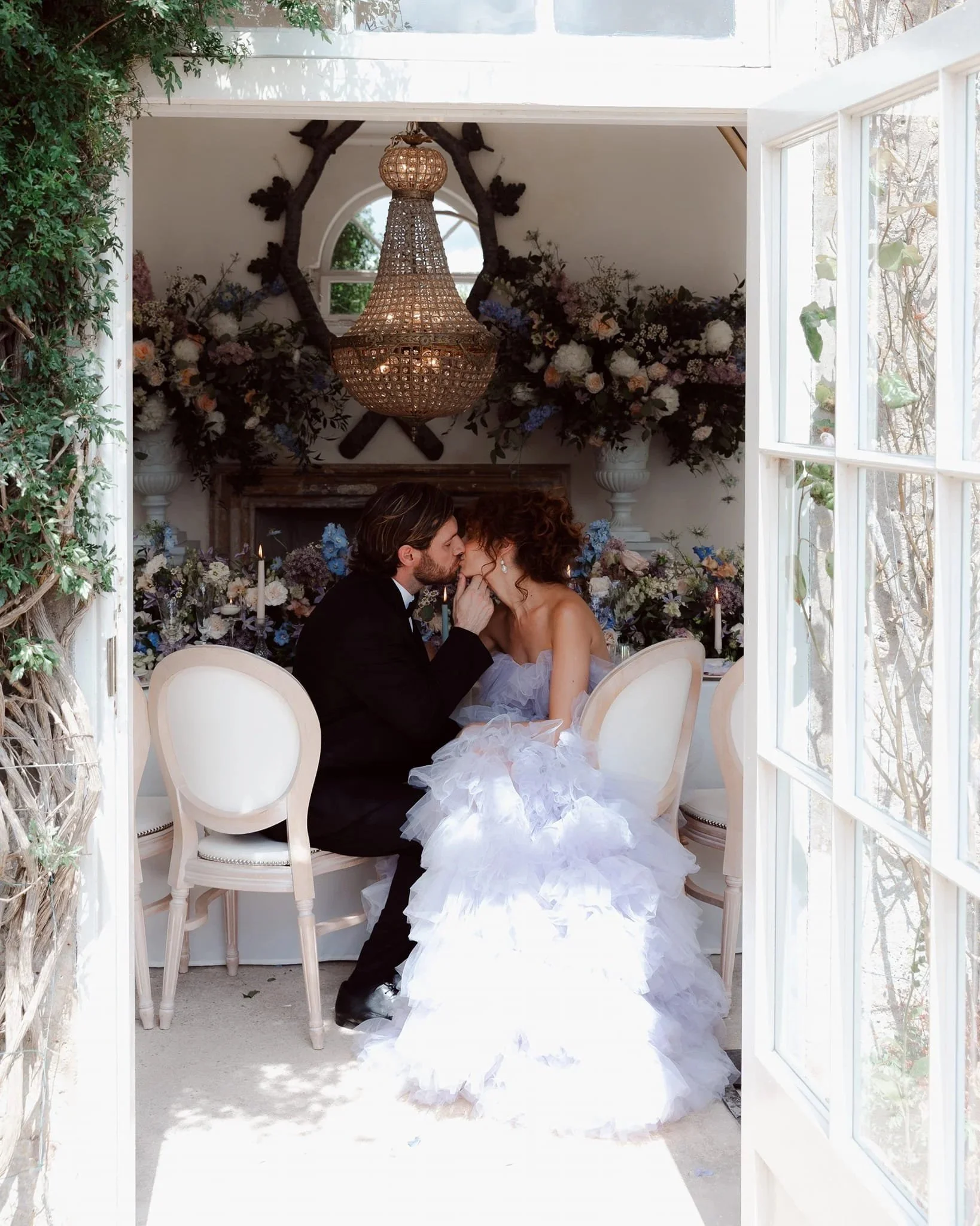 A bride and groom sharing an intimate moment inside a decorated room with a chandelier and floral arrangements, viewed through a window with open doors.