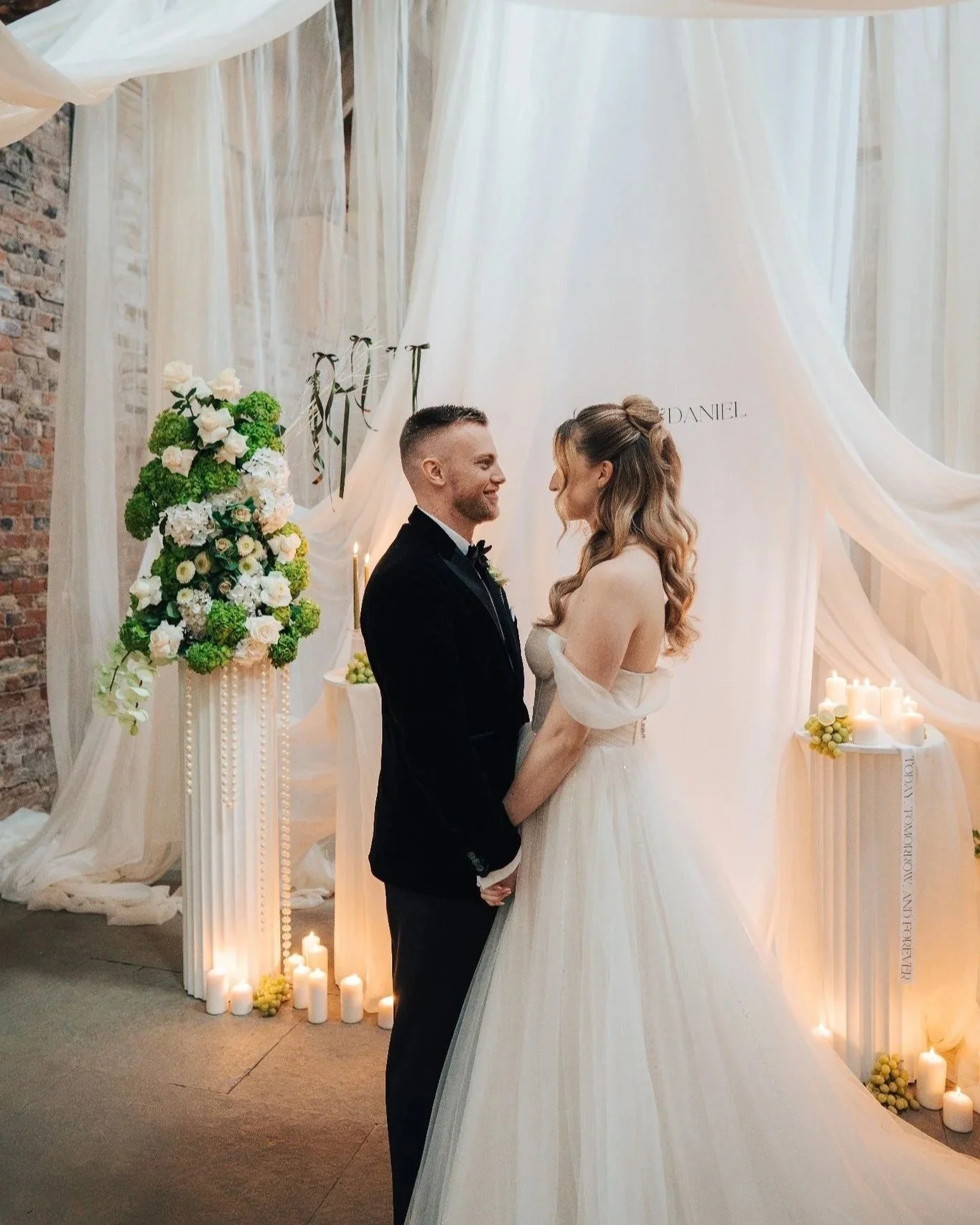 A bride and groom hold hands and smile at each other during their wedding ceremony, with floral arrangements and candles in the background.