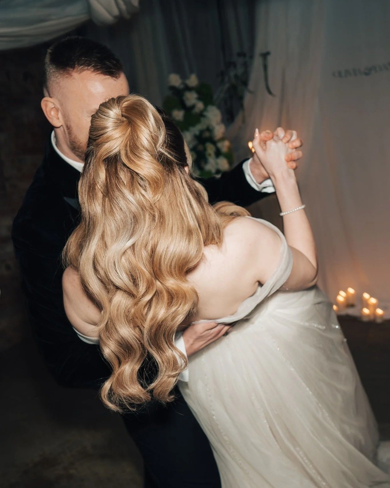 A couple is dancing closely at their wedding, with the woman wearing a strapless white gown and the man in a tuxedo, holding hands and dancing in a dimly lit room with candles and flowers in the background.