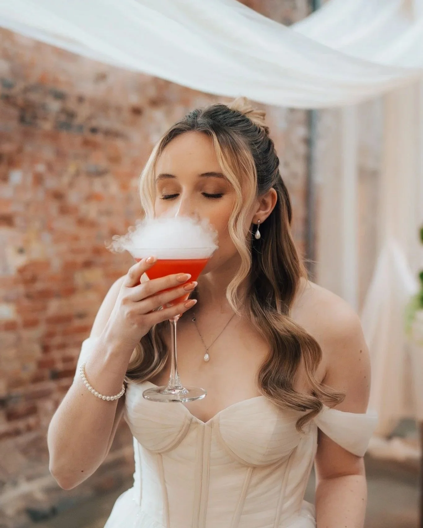 A woman in a white dress and pearl jewelry, with her hair styled in loose curls, holds a cocktail glass filled with a red drink topped with dry ice smoke, in an indoor setting with a brick wall background.