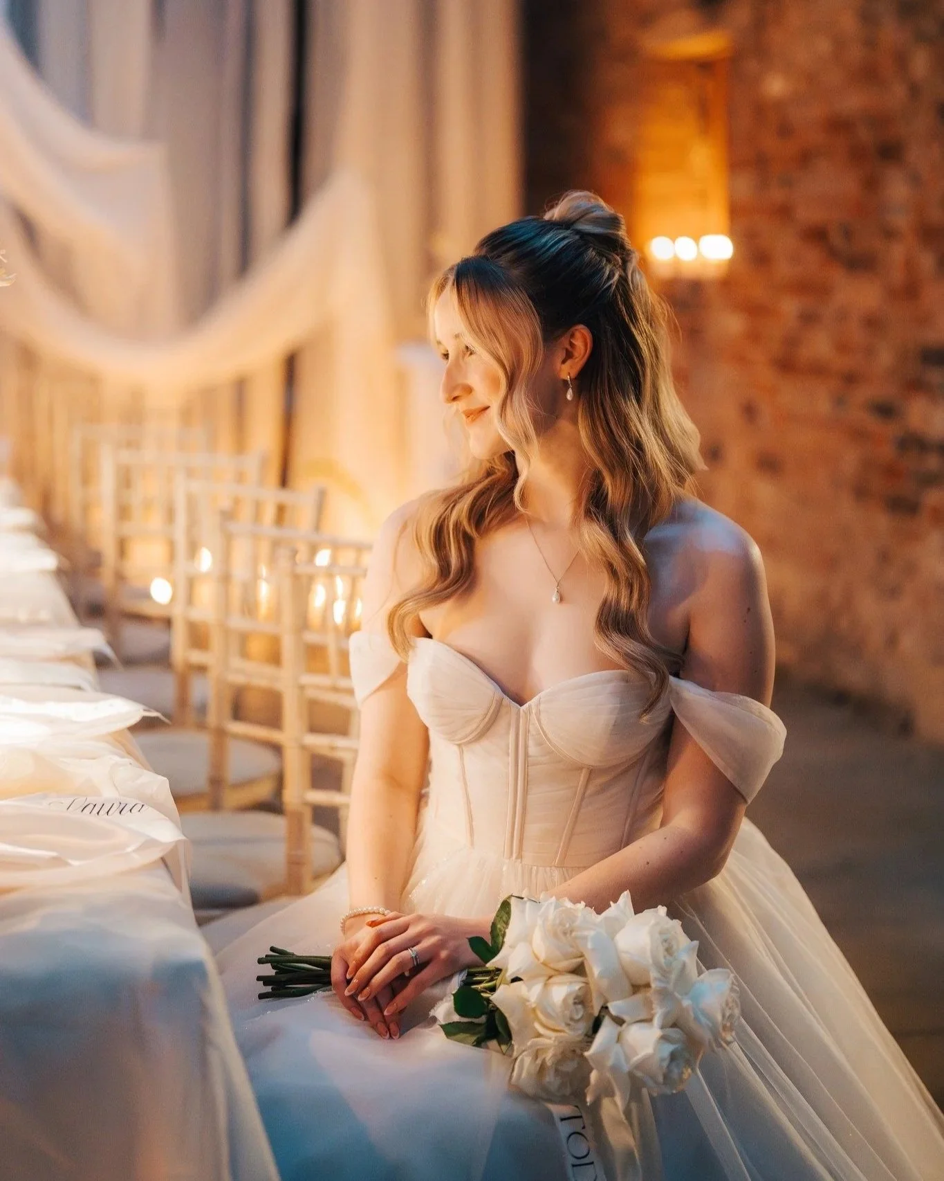 A bride in a strapless wedding gown sitting with a bouquet of white roses, smiling gently with her eyes closed in a warmly lit indoor setting.