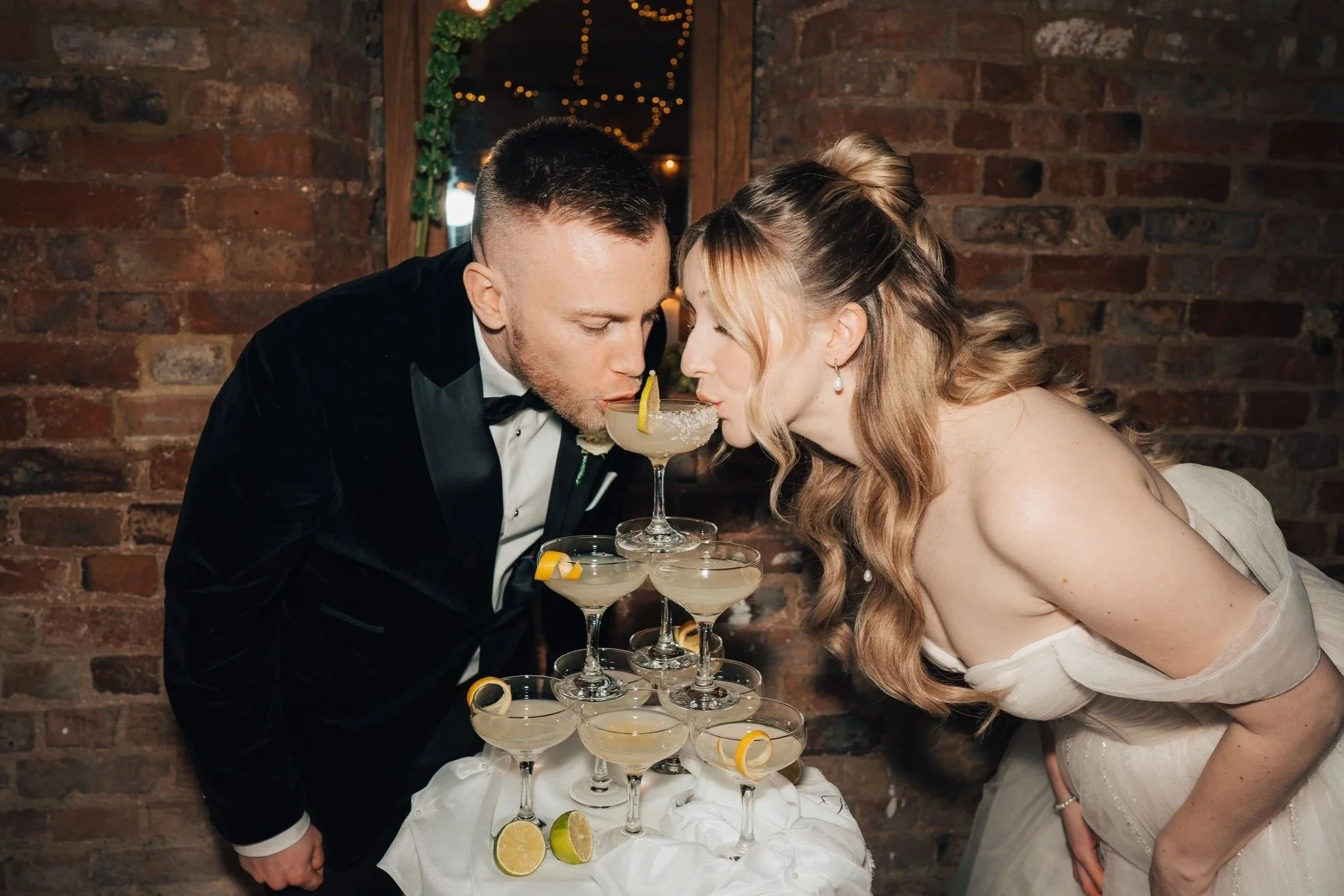 A bride and groom at their wedding, toasting with cocktails balanced on a multi-tiered stand, forming a wedding cake, in front of an exposed brick wall.