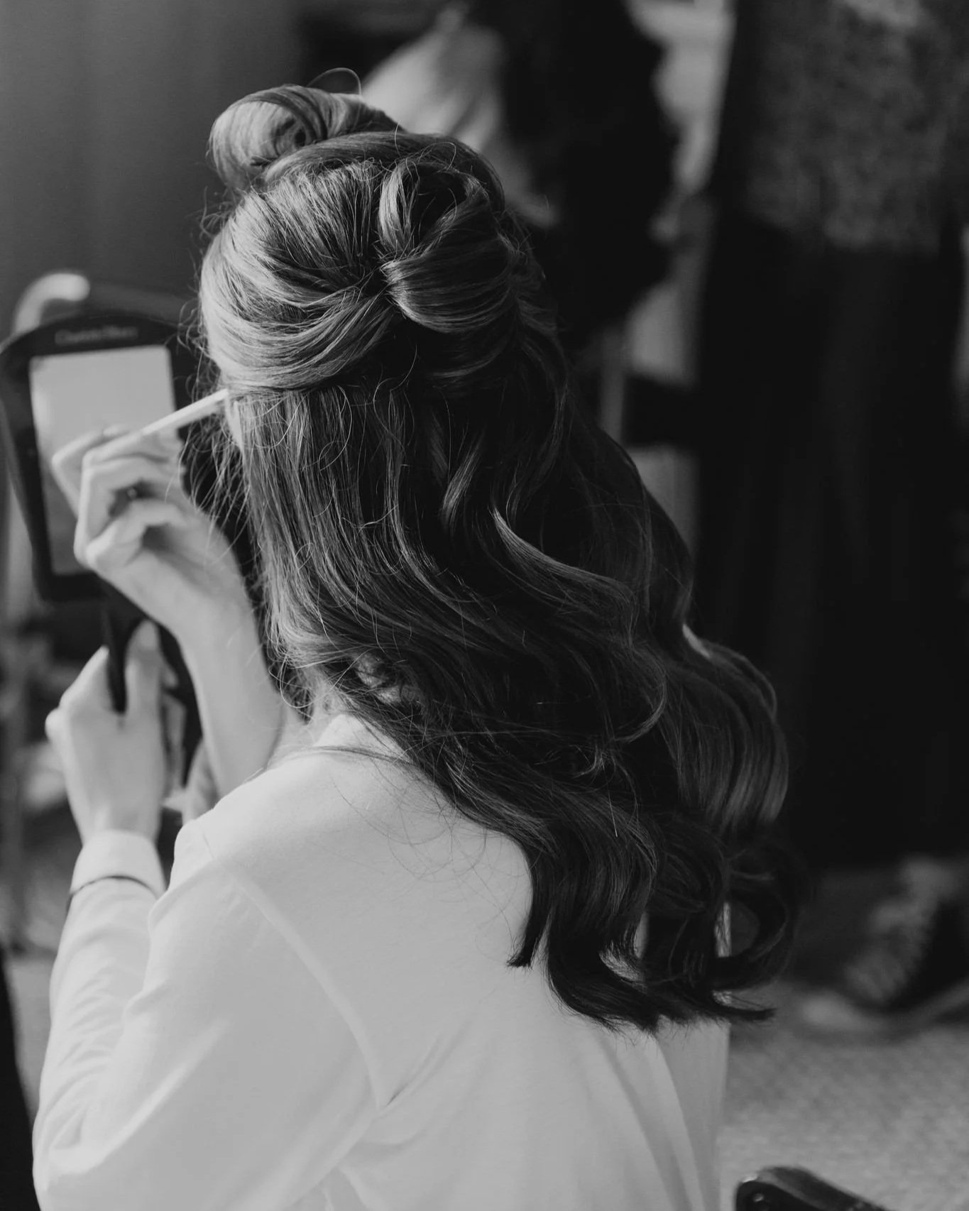A woman with styled, curled hair is applying makeup or skincare using a small mirror, sitting indoors.