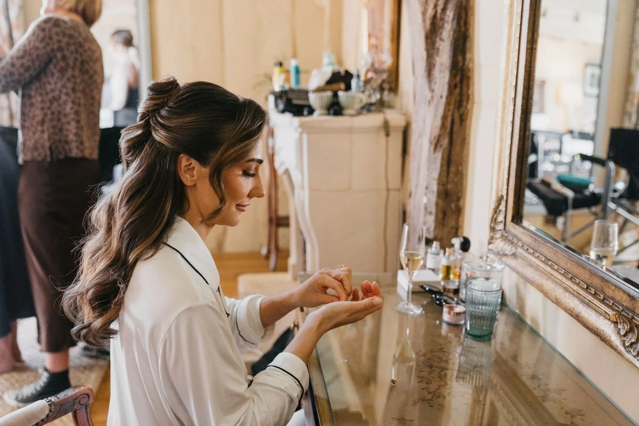 Woman in white robe applying makeup in front of mirror, with friends in background