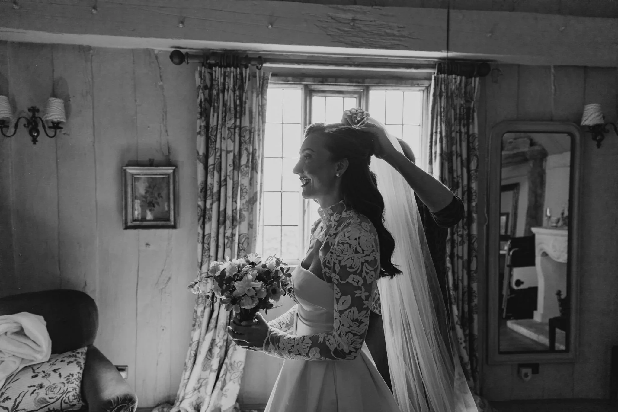 A bride in a wedding dress holding a bouquet, smiling while someone helps her with her veil in a cozy, vintage room with a window, curtains, and a mirror.