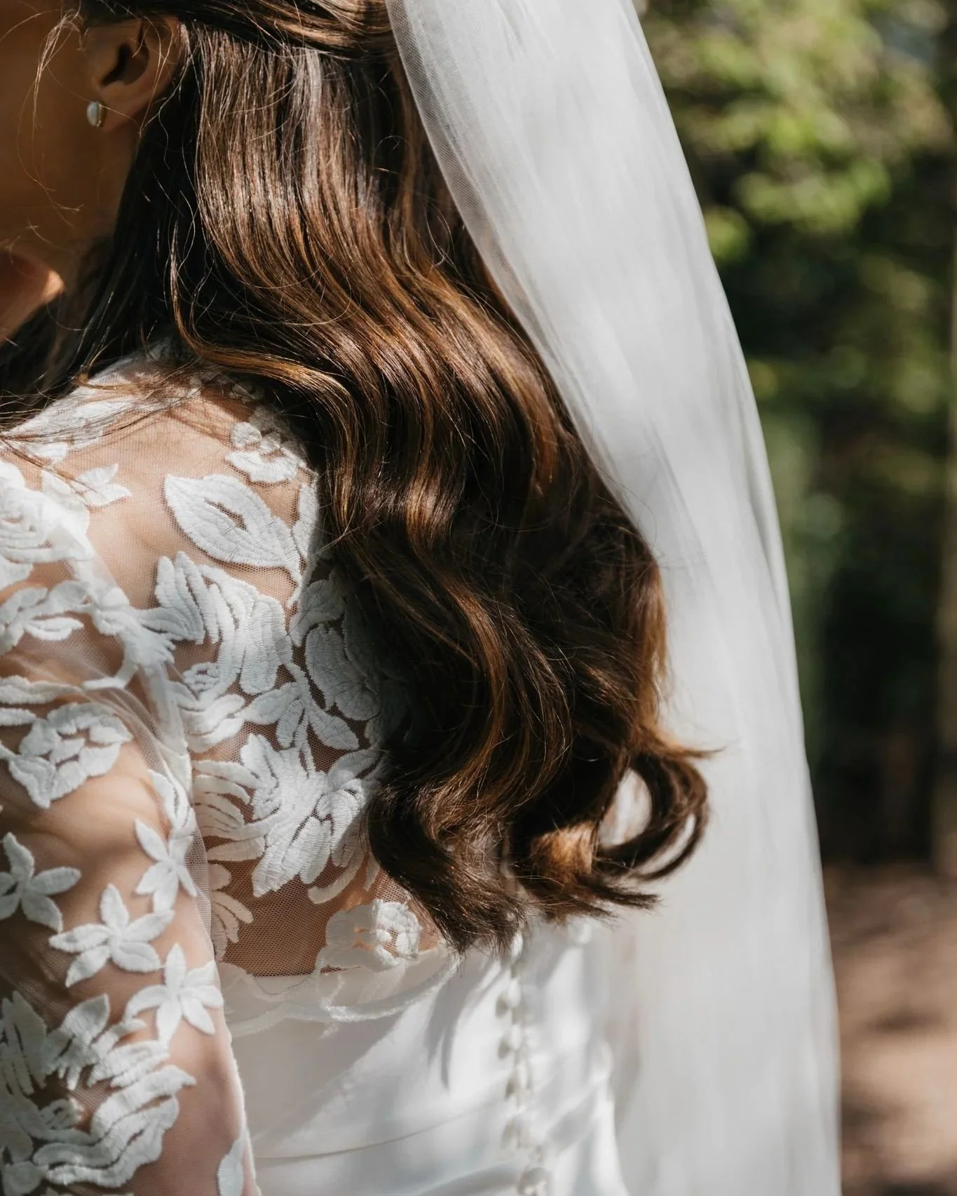 Close-up of a bride with long, wavy brunette hair wearing a white wedding dress with lace floral embroidery and a veil.