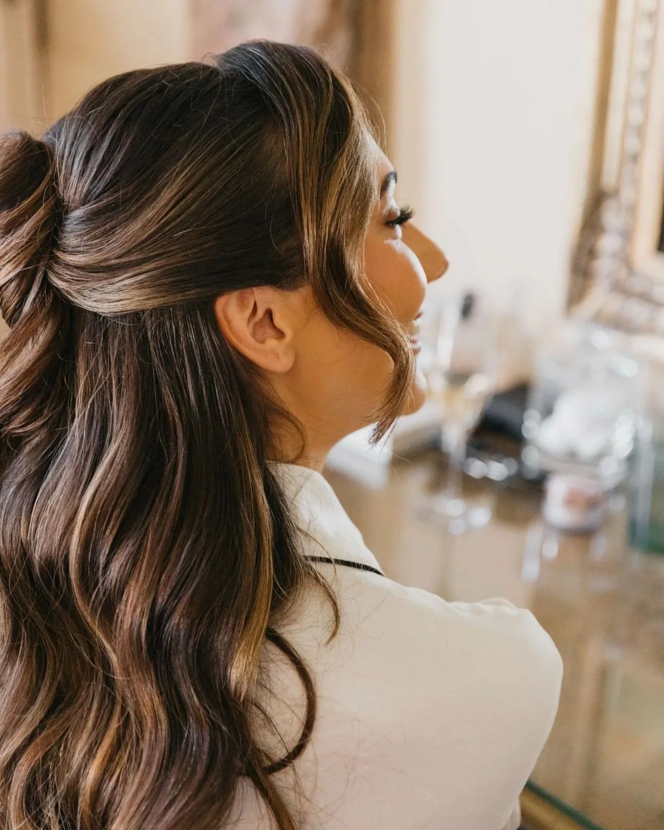 A woman with long, wavy brown hair and highlights, seen from the side, sitting in a room with a mirror and makeup items in the background.