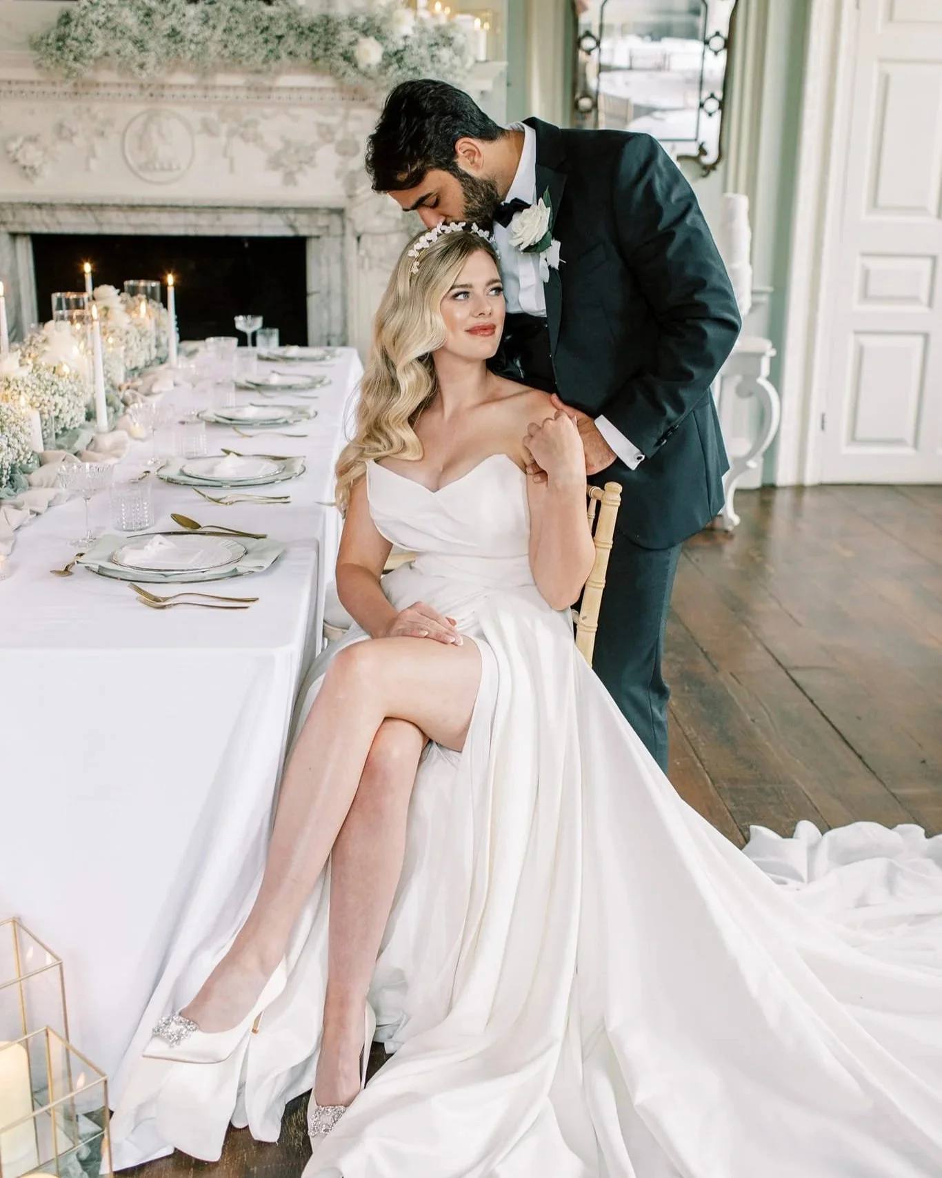 Bride sitting on a chair wearing a white wedding gown with a train, and a groom standing behind her with a wedding boutonniere, in a decorated wedding venue with a long table set for a celebration.
