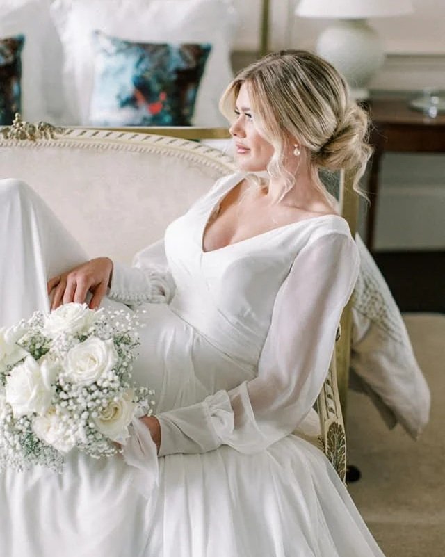 A bride in a white wedding dress sitting on a vintage sofa, holding a bouquet of white roses and baby's breath, with decorated pillows in the background.
