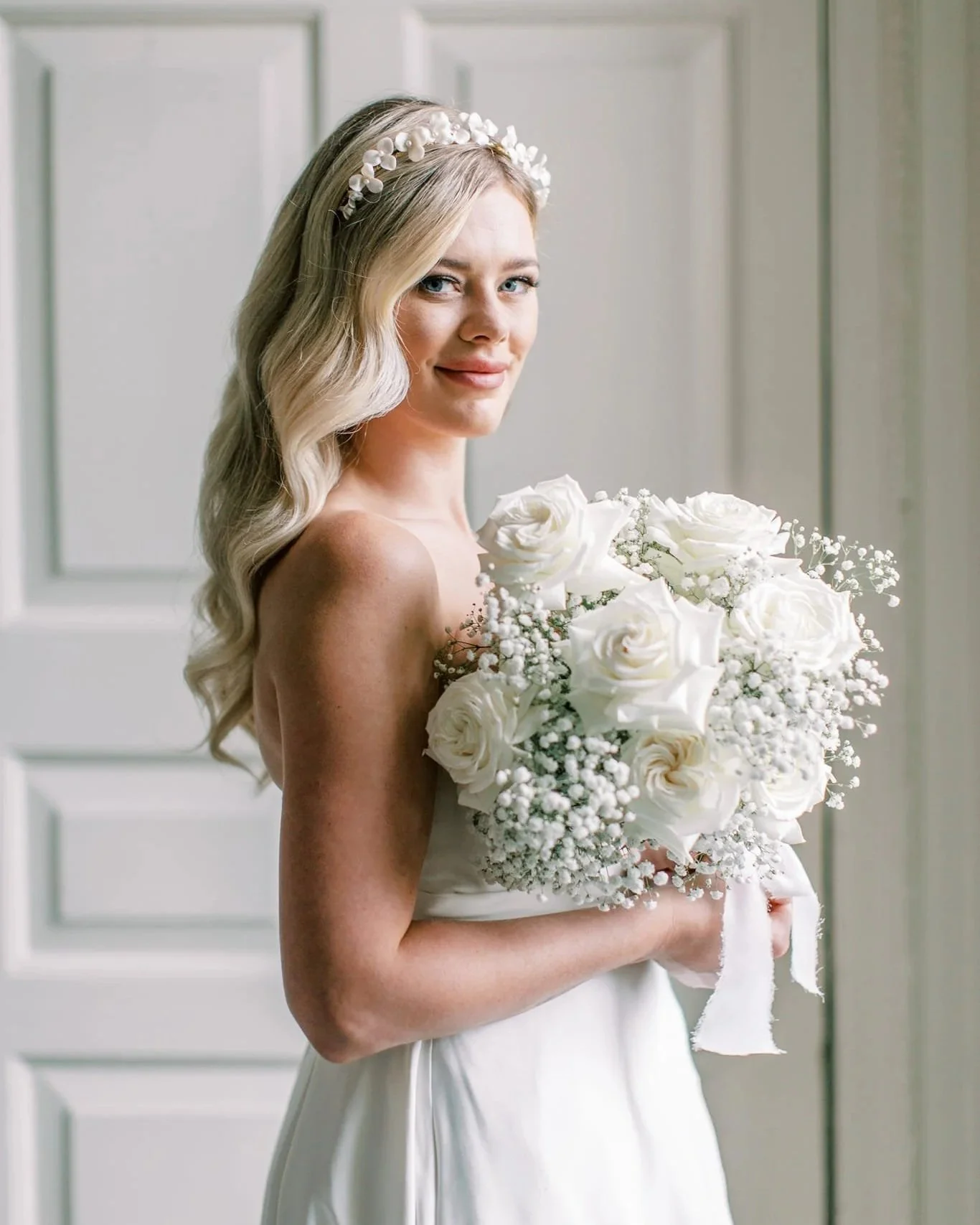 A bride with long blonde hair, wearing a floral headband, holding a bouquet of white roses and baby's breath, standing in front of a white door.