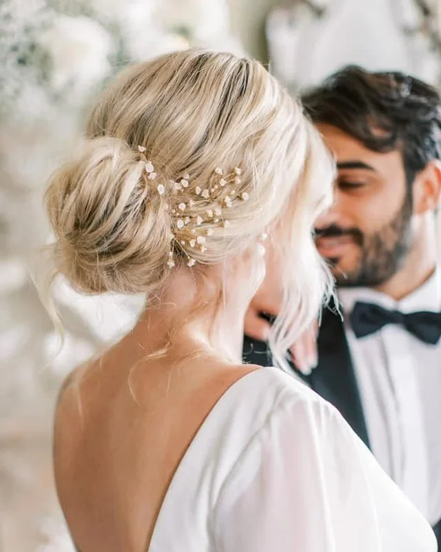 Bride and groom sharing a close, intimate moment at their wedding, with the bride's elegant updo hairstyle decorated with small white flowers and pearl accents, and the groom wearing a tuxedo with a bow tie.