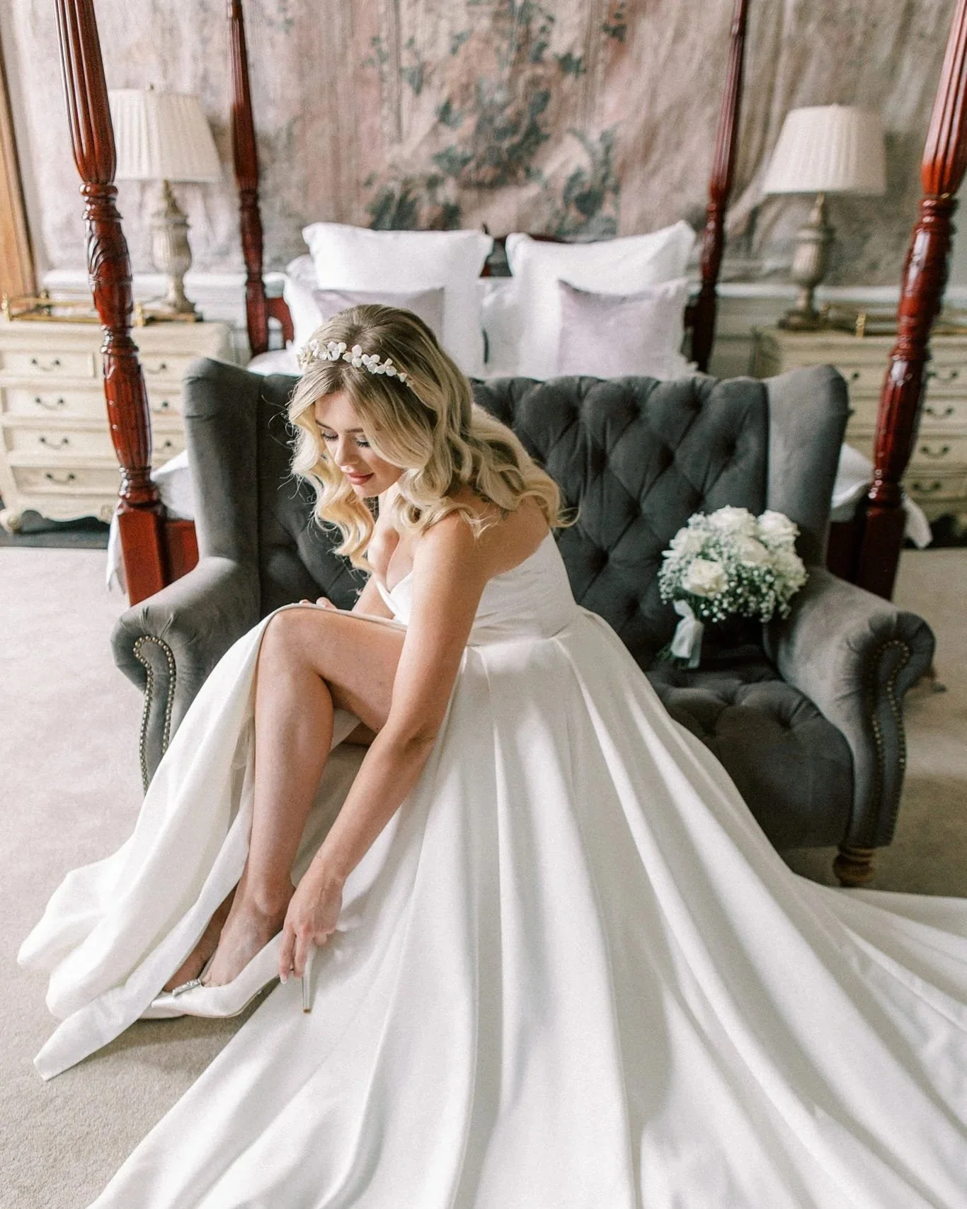 A bride in a white wedding gown sitting on a gray velvet sofa, putting on her white high heels, in a richly decorated bedroom with a four-poster bed, white bedding, and floral arrangements.