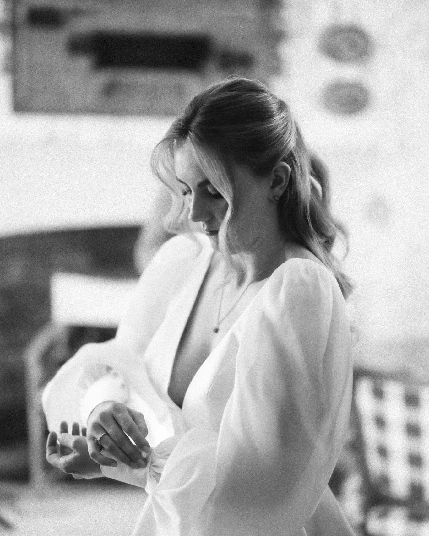 A woman with wavy hair looking down, wearing a light-colored dress, standing in an indoor setting.