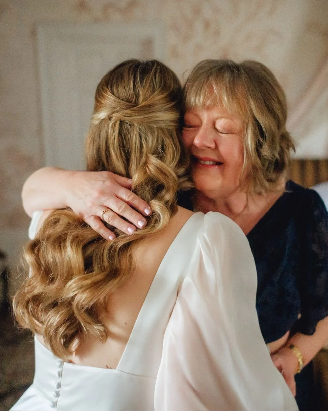 A woman with blonde hair hugging a younger woman with long, wavy, light brown hair, both smiling and showing affection.