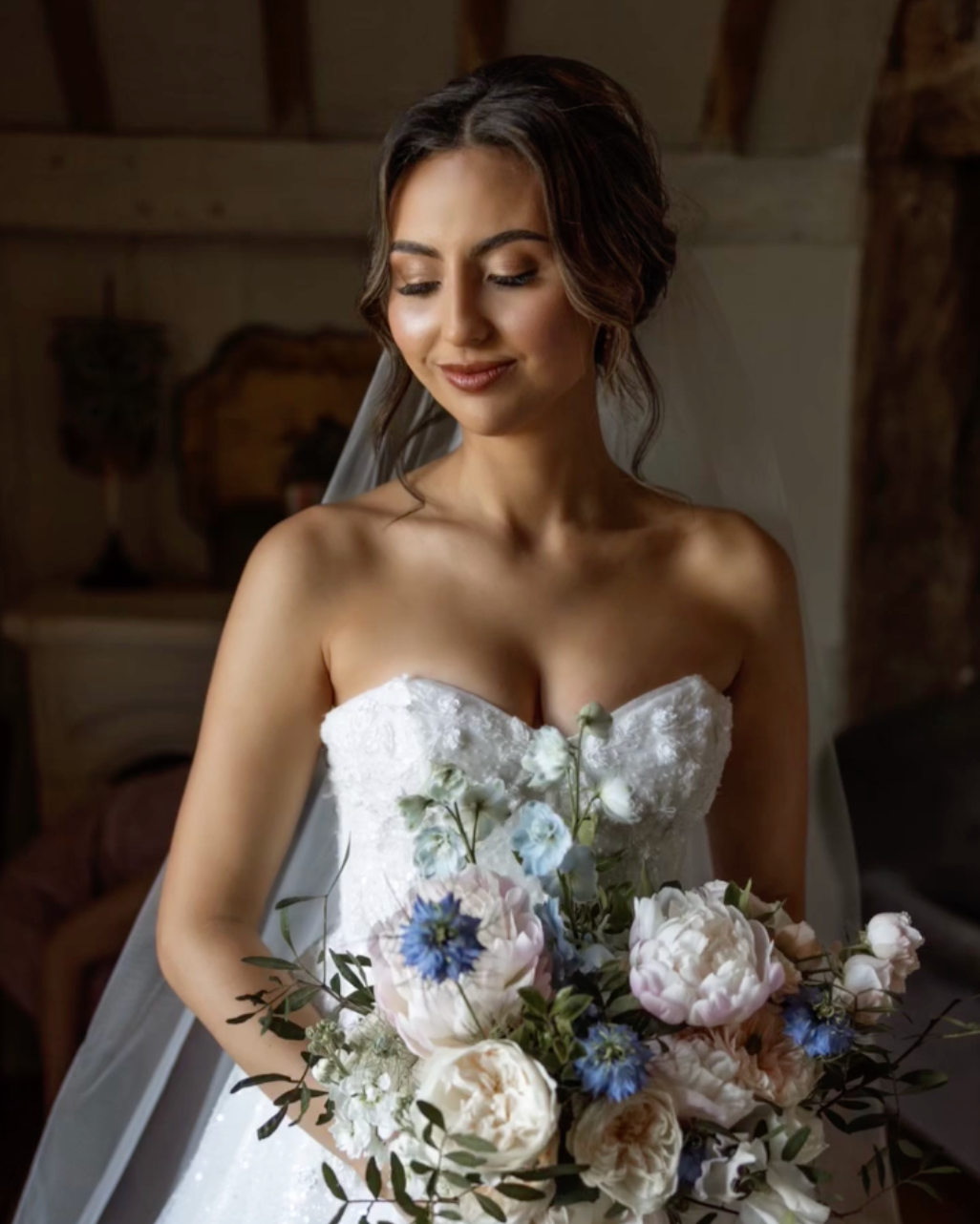 A bride in a strapless white wedding dress holding a bouquet of pink, white, and blue flowers, with a soft smile and eyes closed, in a rustic indoor setting.