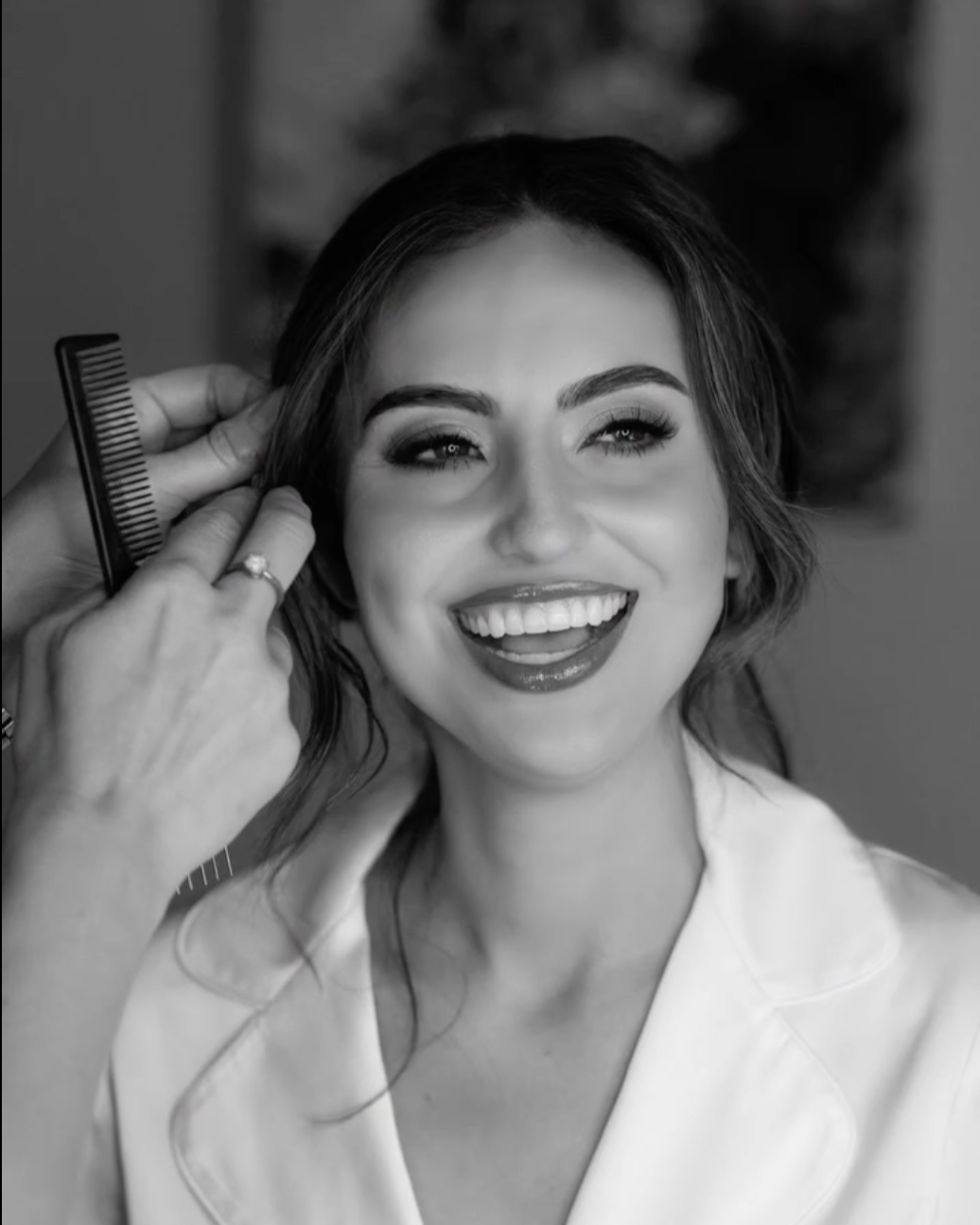 A woman with a bright smile getting her hair styled, with a stylist holding a comb near her head.