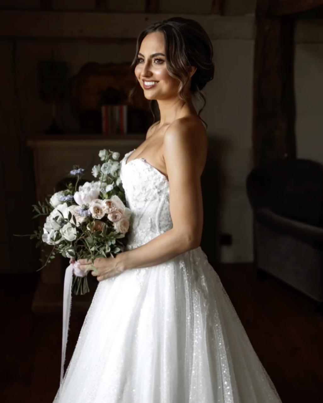 A bride in a strapless white wedding dress holding a bouquet of flowers, smiling inside a rustic wooden room.
