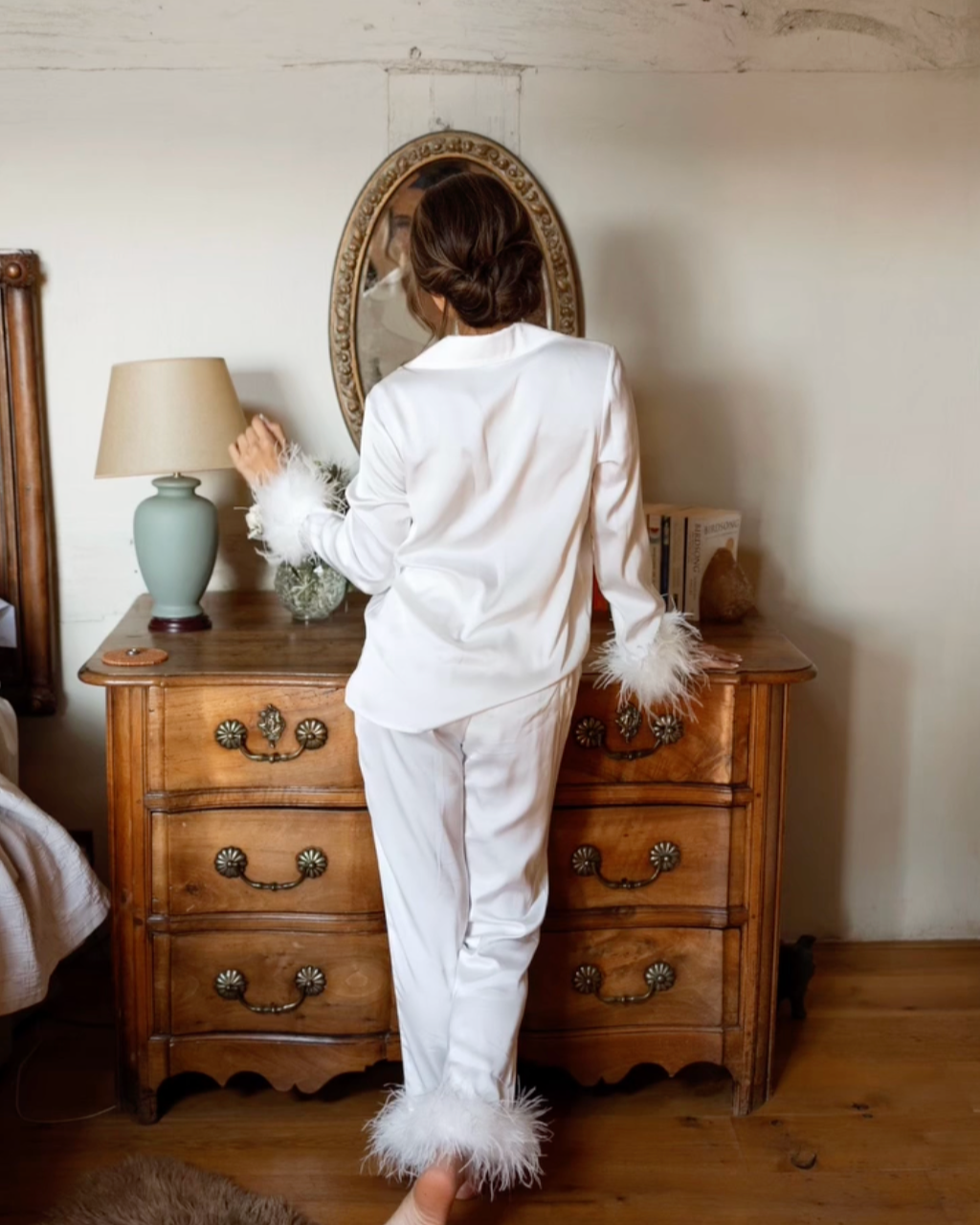 A woman in white satin pajamas with feather trim on the sleeves and pant cuffs standing in a bedroom, looking into a mirror on a wooden dresser.