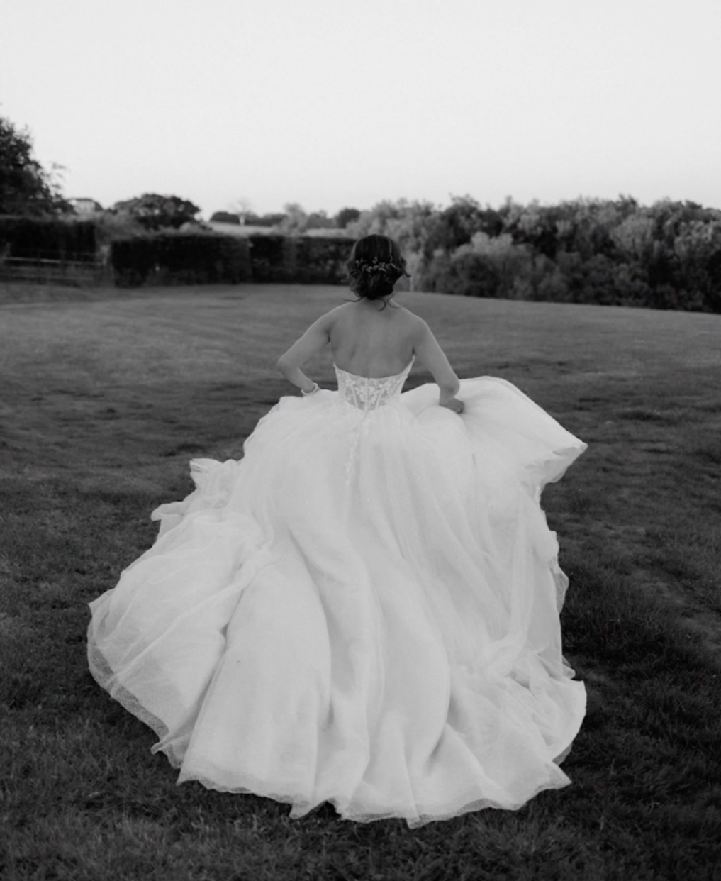A woman in a white wedding dress walking on a grassy field, viewed from behind.