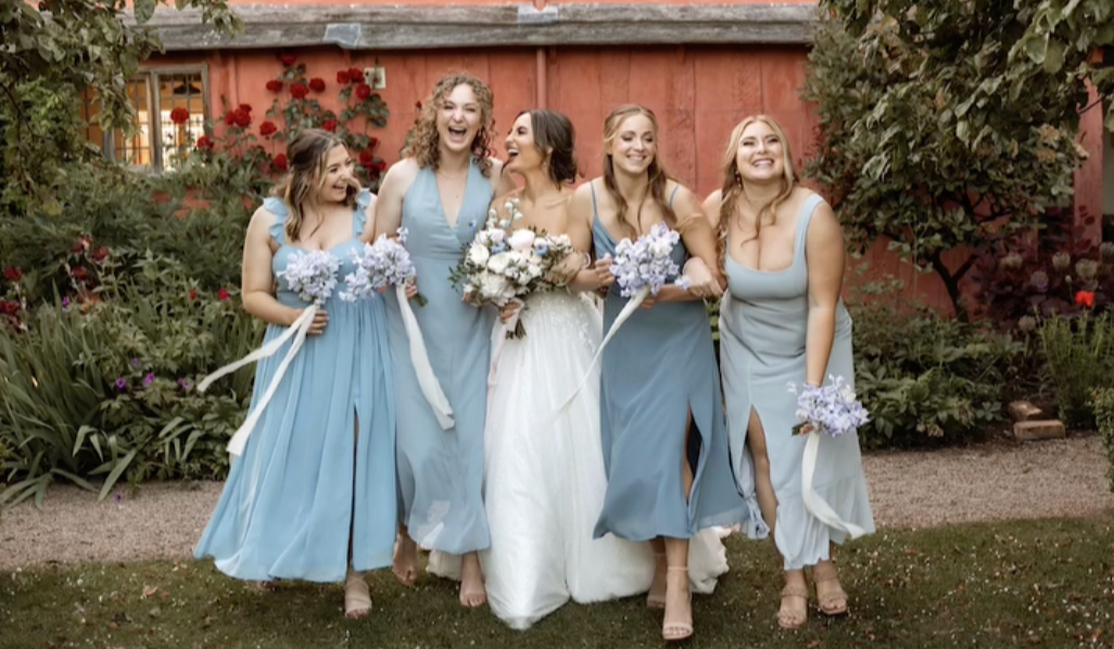A bride and five bridesmaids laughing and walking in a garden. The bride holds a bouquet, and the bridesmaids hold smaller bouquets. They are wearing matching blue dresses and beige heels.