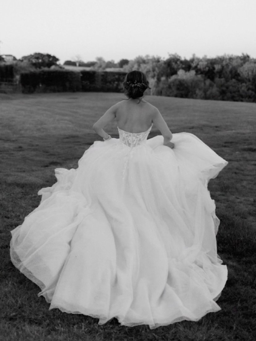 A woman in a white wedding dress walking on a grassy field, viewed from behind.