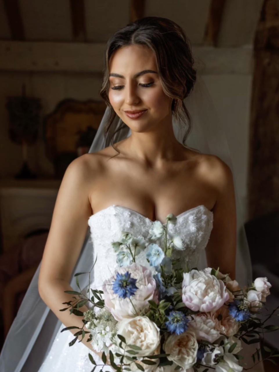 A bride in a strapless white wedding dress holding a bouquet of pink, white, and blue flowers, with a soft smile and eyes closed, in a rustic indoor setting.