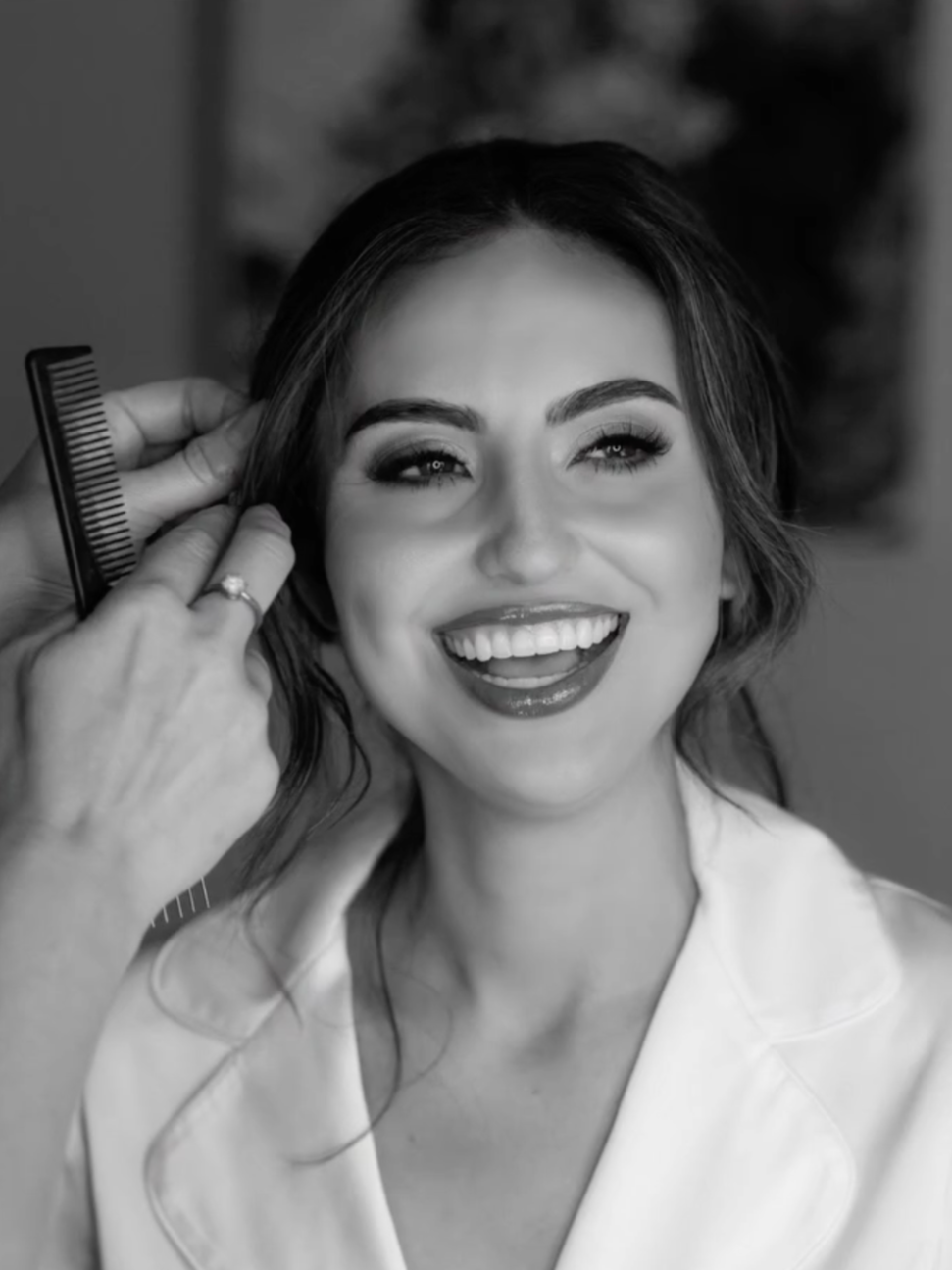 A woman with a bright smile getting her hair styled, with a stylist holding a comb near her head.