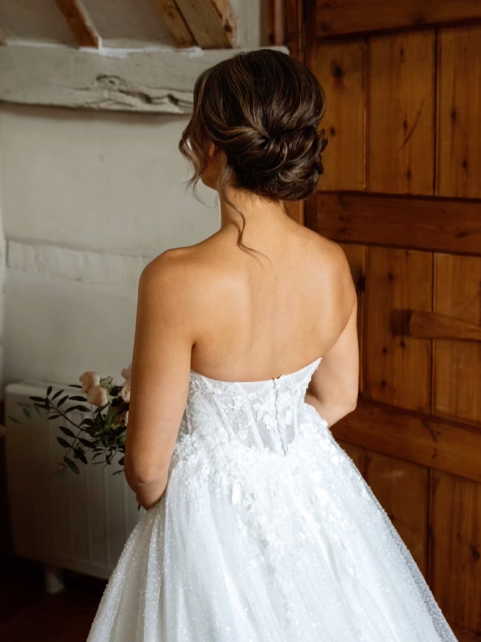 A woman in a white strapless wedding dress with lace detailing, standing with her back to the camera in a rustic room with wooden walls and a white stone fireplace.