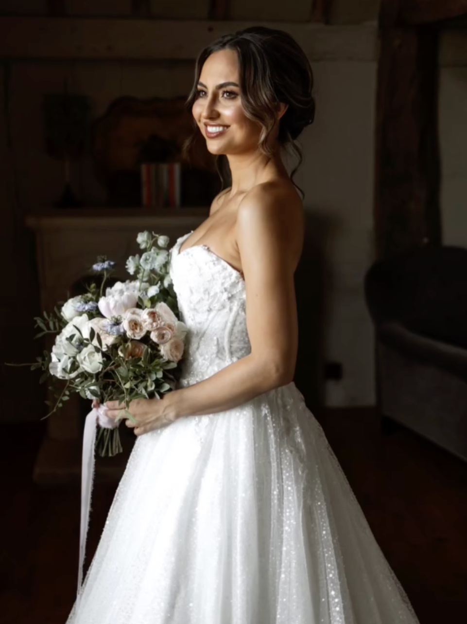A bride in a strapless white wedding dress holding a bouquet of flowers, smiling inside a rustic wooden room.