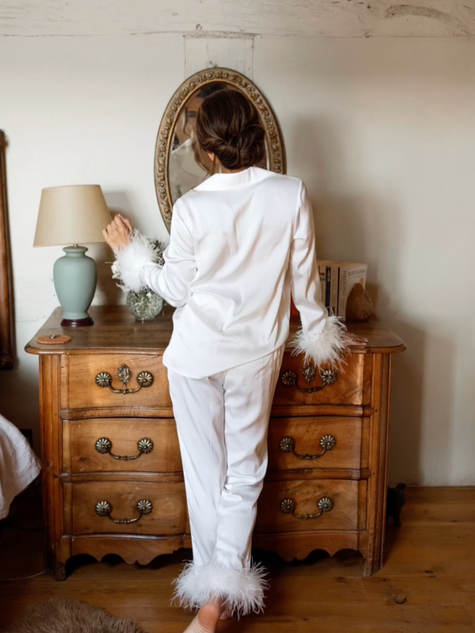A woman in white satin pajamas with feather trim on the sleeves and pant cuffs standing in a bedroom, looking into a mirror on a wooden dresser.