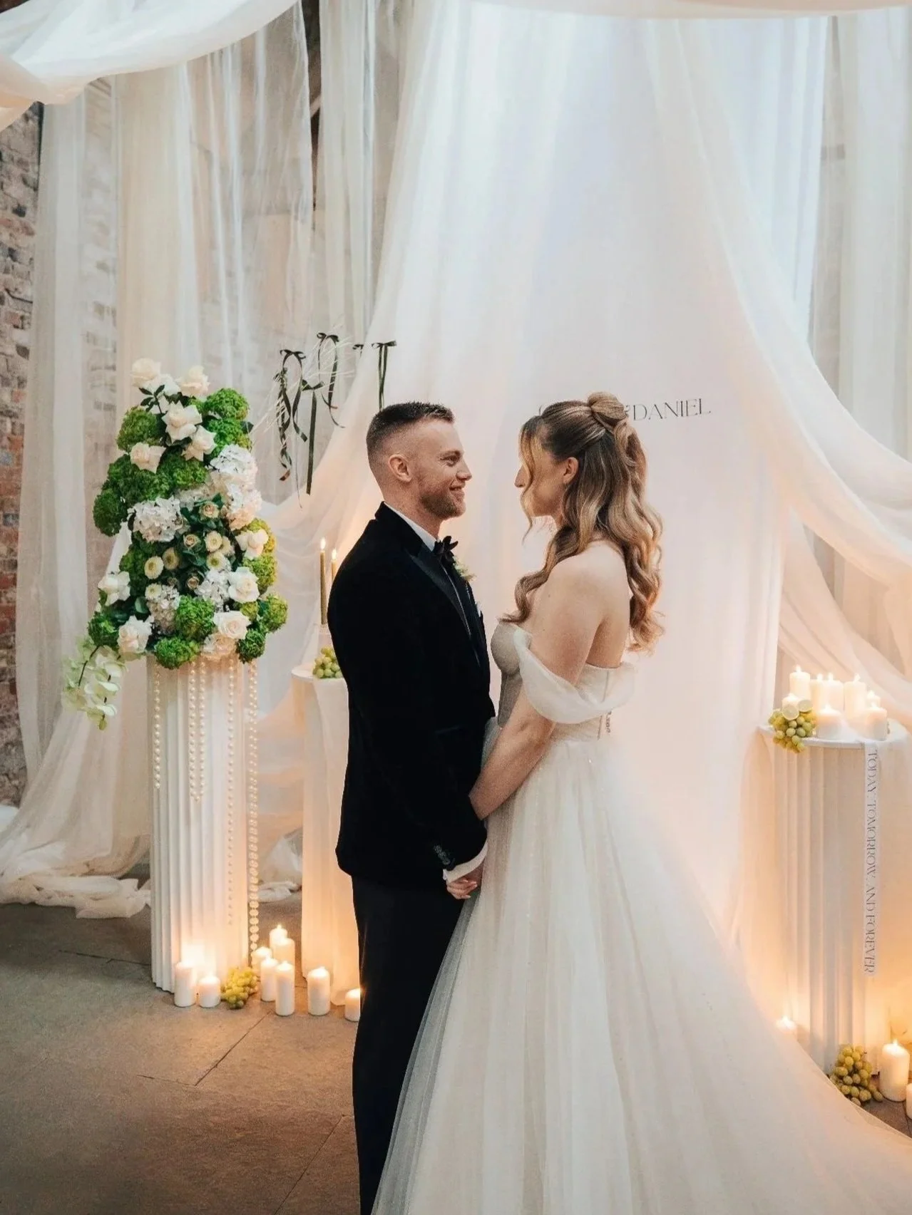 A bride and groom hold hands and smile at each other during their wedding ceremony, with floral arrangements and candles in the background.