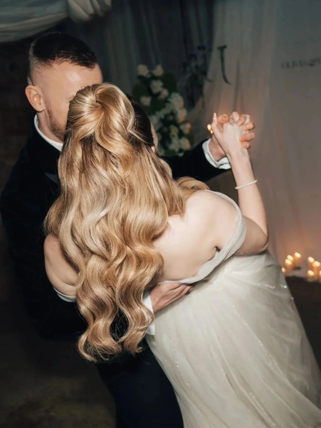 A couple is dancing closely at their wedding, with the woman wearing a strapless white gown and the man in a tuxedo, holding hands and dancing in a dimly lit room with candles and flowers in the background.