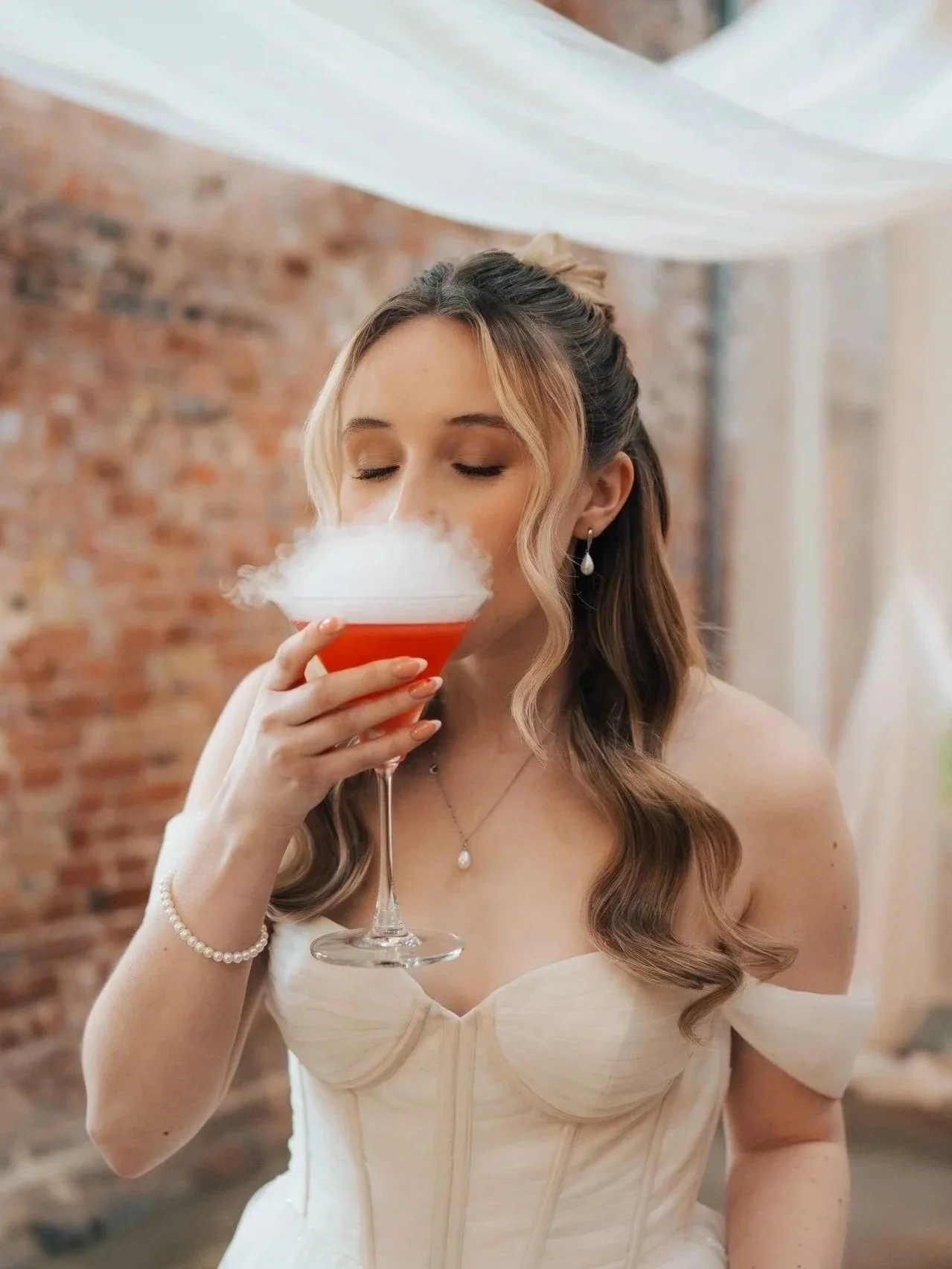 A woman in a white dress and pearl jewelry, with her hair styled in loose curls, holds a cocktail glass filled with a red drink topped with dry ice smoke, in an indoor setting with a brick wall background.