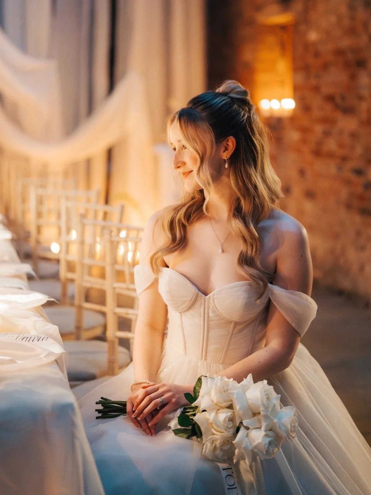 A bride in a strapless wedding gown sitting with a bouquet of white roses, smiling gently with her eyes closed in a warmly lit indoor setting.