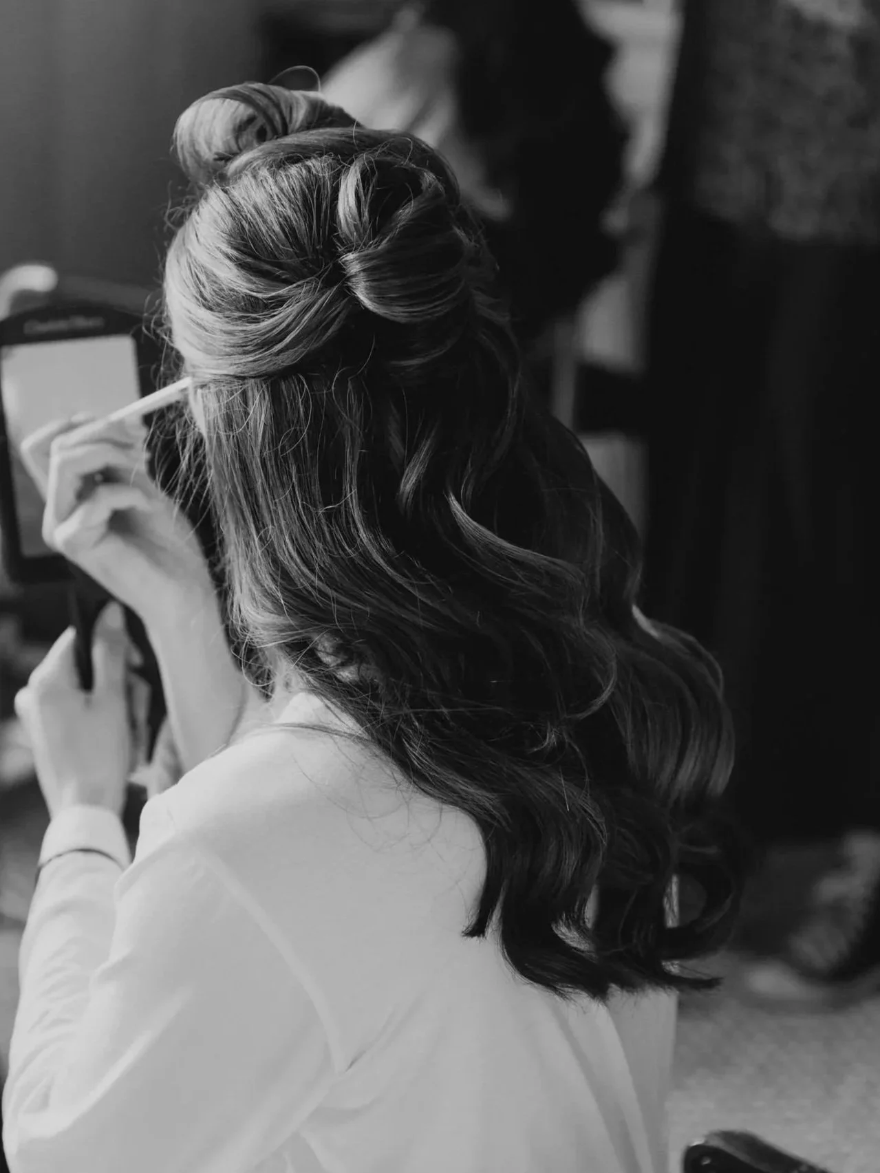 A woman with styled, curled hair is applying makeup or skincare using a small mirror, sitting indoors.