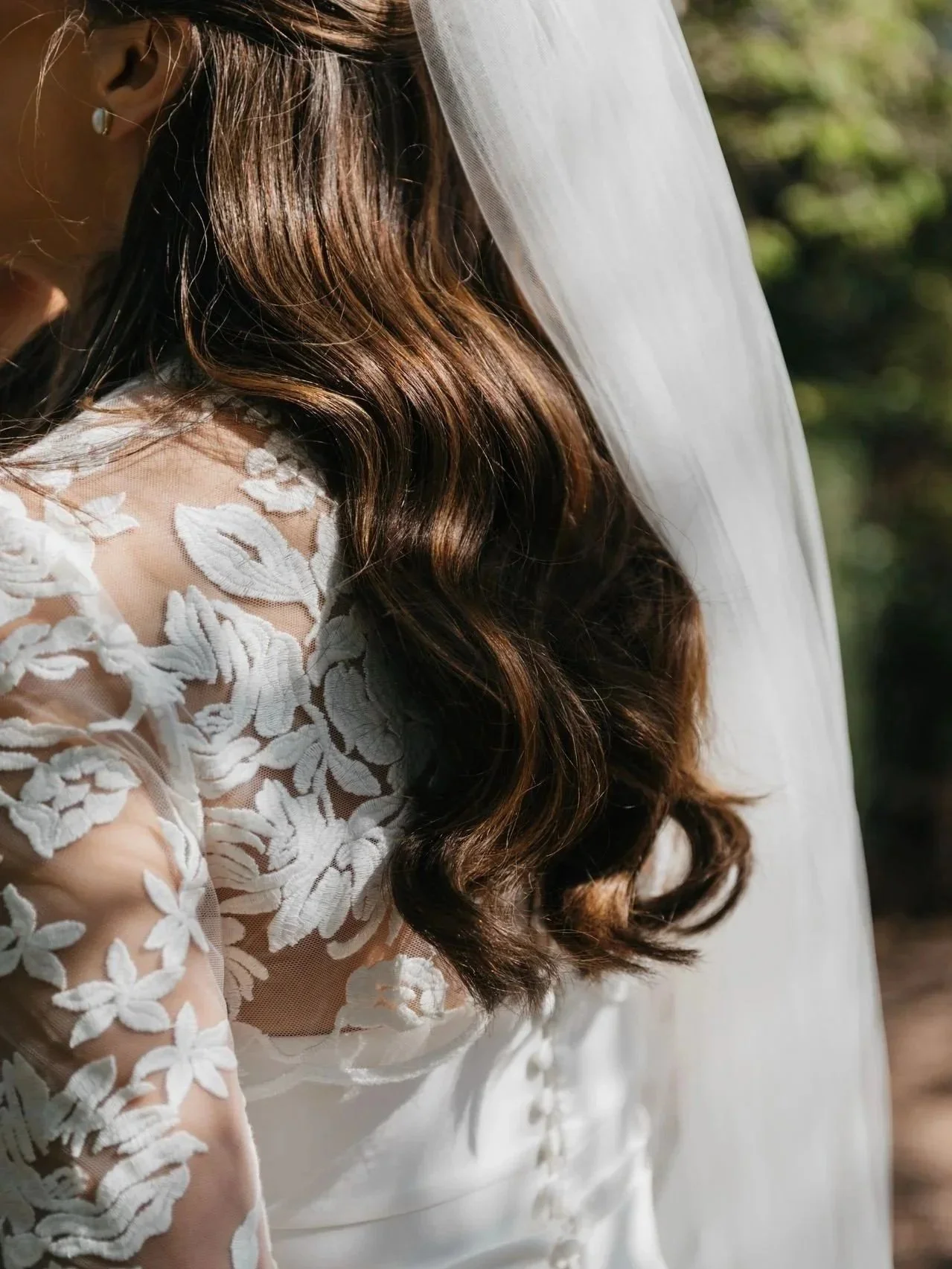 Close-up of a bride with long, wavy brunette hair wearing a white wedding dress with lace floral embroidery and a veil.