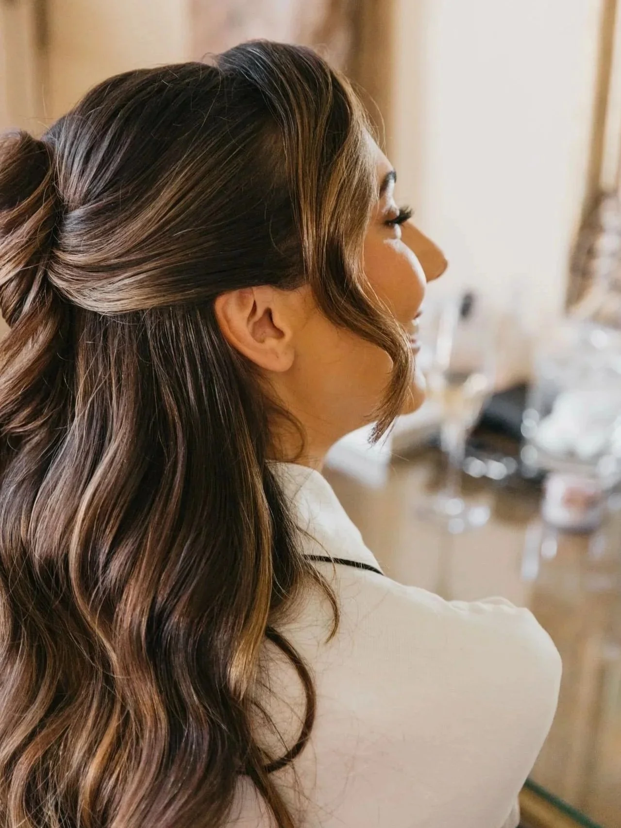 A woman with long, wavy brown hair and highlights, seen from the side, sitting in a room with a mirror and makeup items in the background.