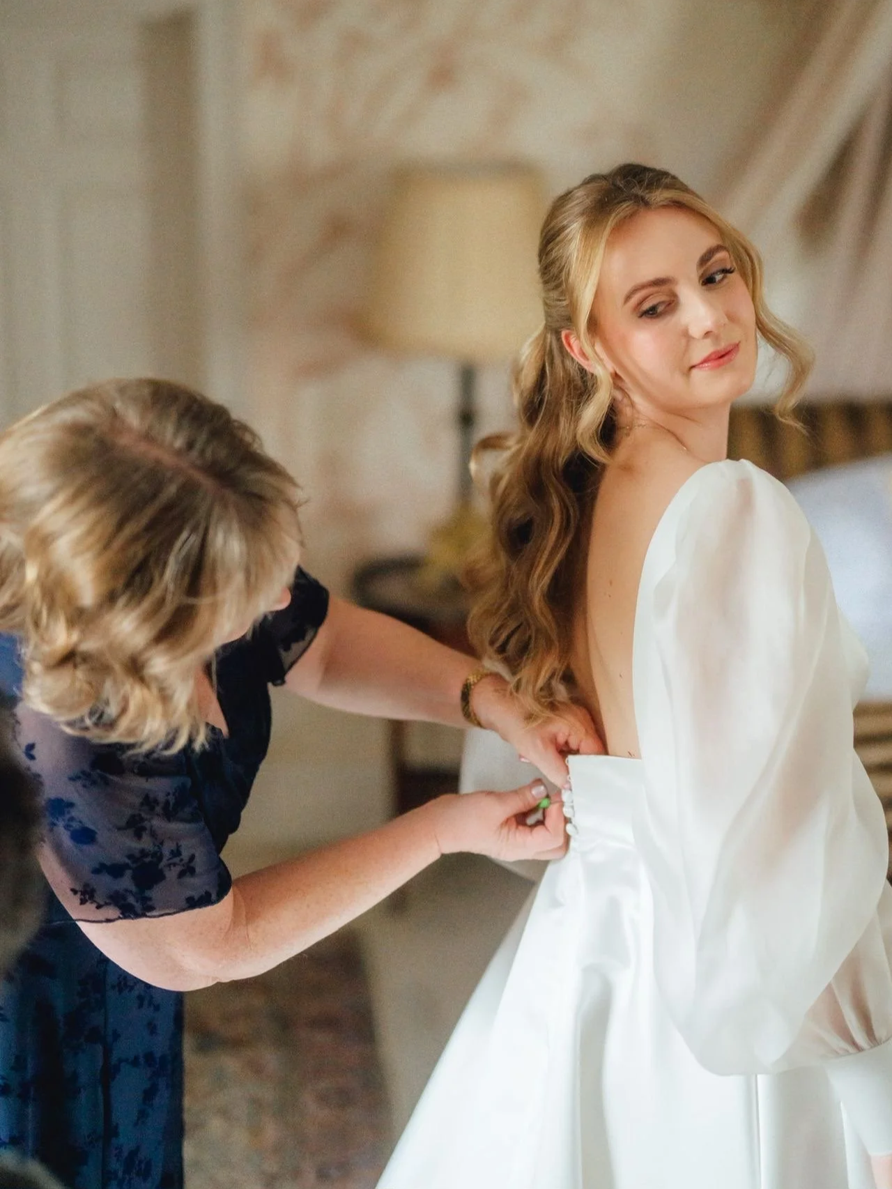 A bride with long, wavy hair in a white wedding dress is assisted by a woman in a dark floral dress as she buttons up the back of her dress in a bedroom.