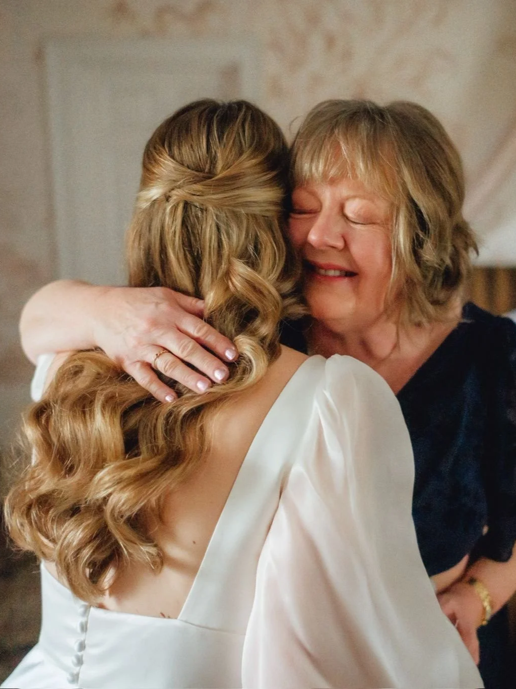 A woman with blonde hair hugging a younger woman with long, wavy, light brown hair, both smiling and showing affection.