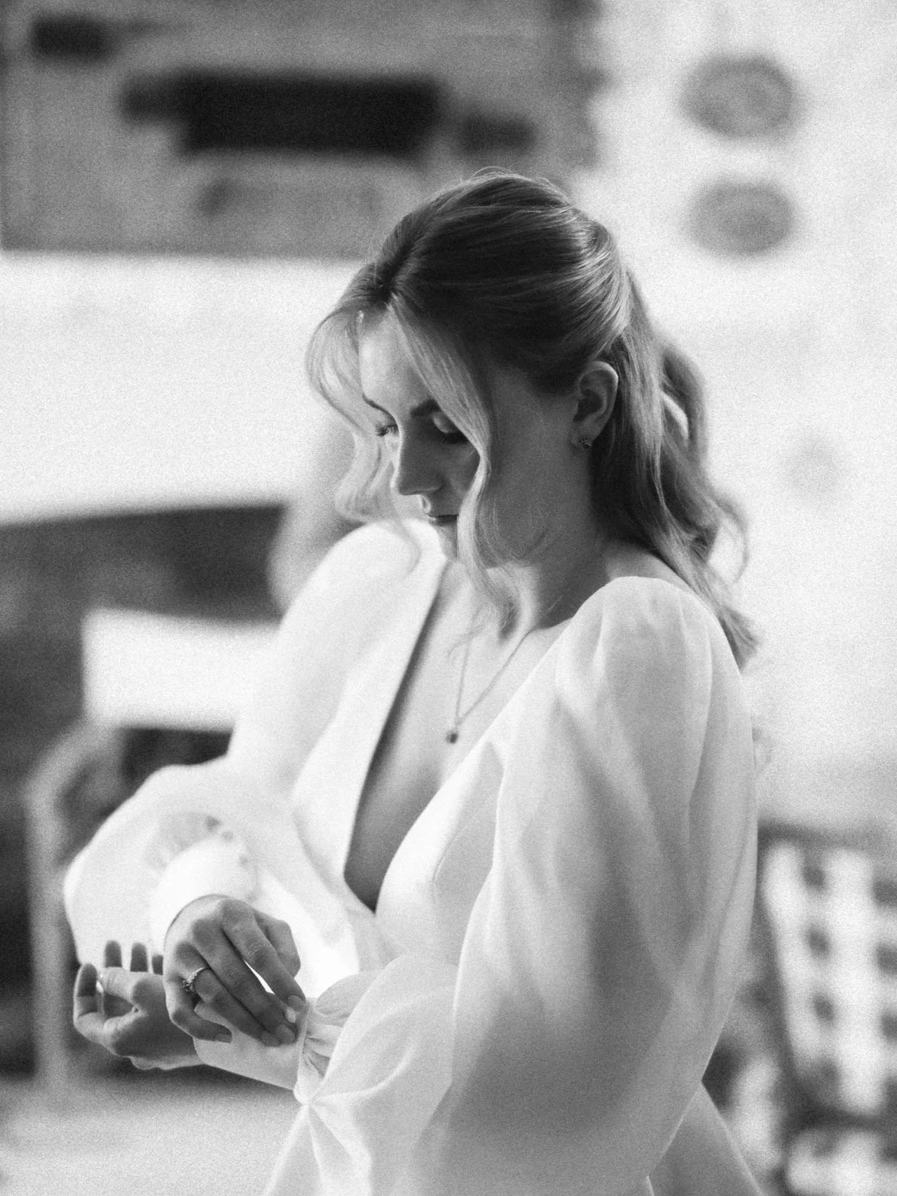 A woman with wavy hair looking down, wearing a light-colored dress, standing in an indoor setting.