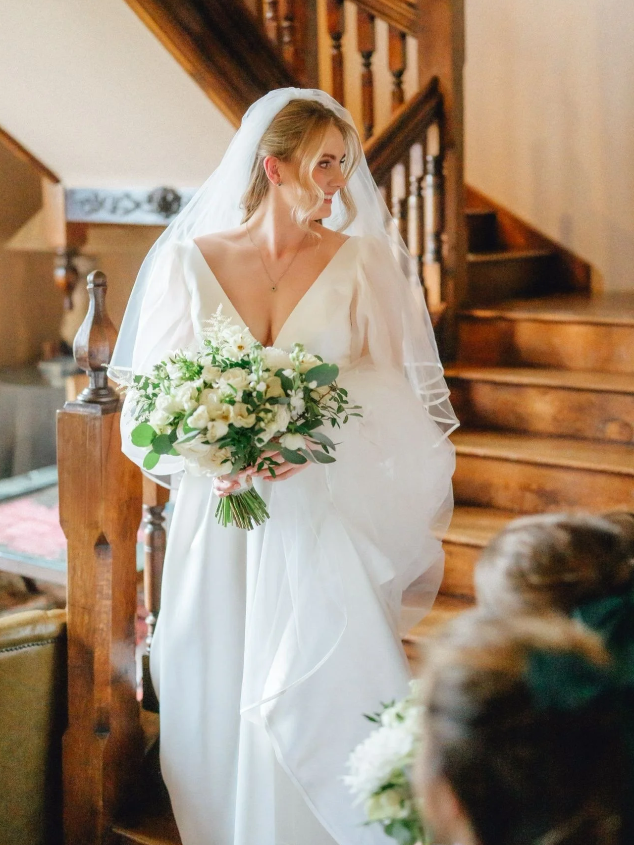 A bride in a white wedding dress holding a bouquet of white flowers and greenery, standing indoors near a wooden staircase.