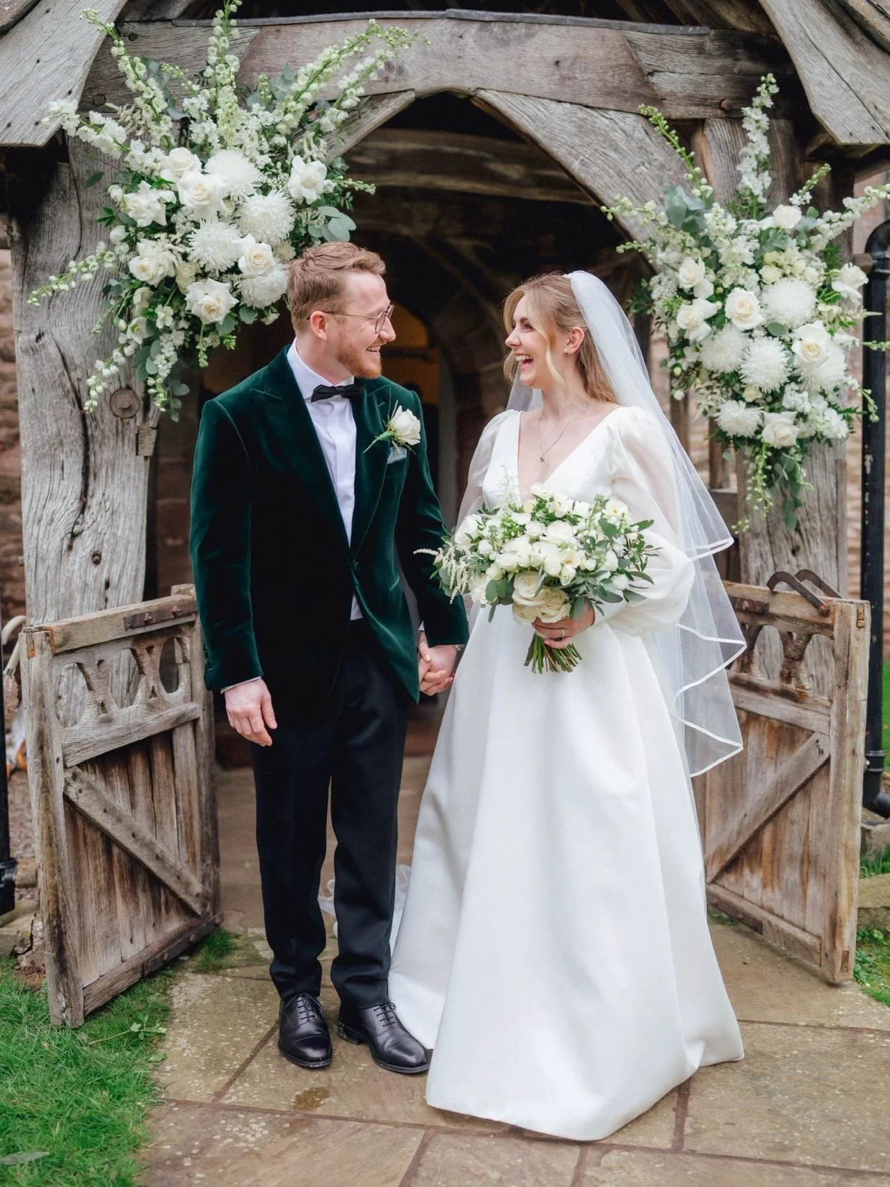 A bride and groom stand in front of a rustic wooden arch decorated with white flowers, holding hands, smiling, and looking at each other during their wedding ceremony.