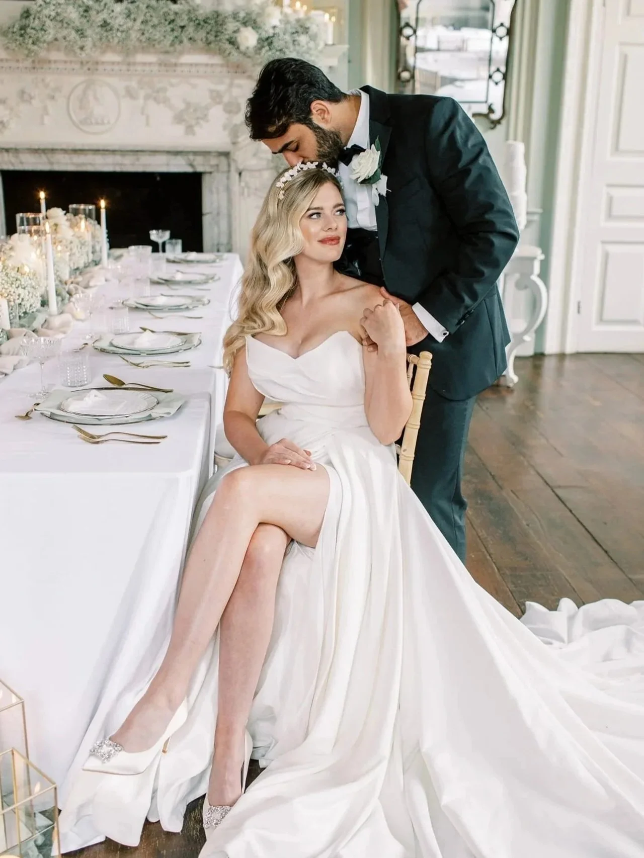 Bride sitting on a chair wearing a white wedding gown with a train, and a groom standing behind her with a wedding boutonniere, in a decorated wedding venue with a long table set for a celebration.