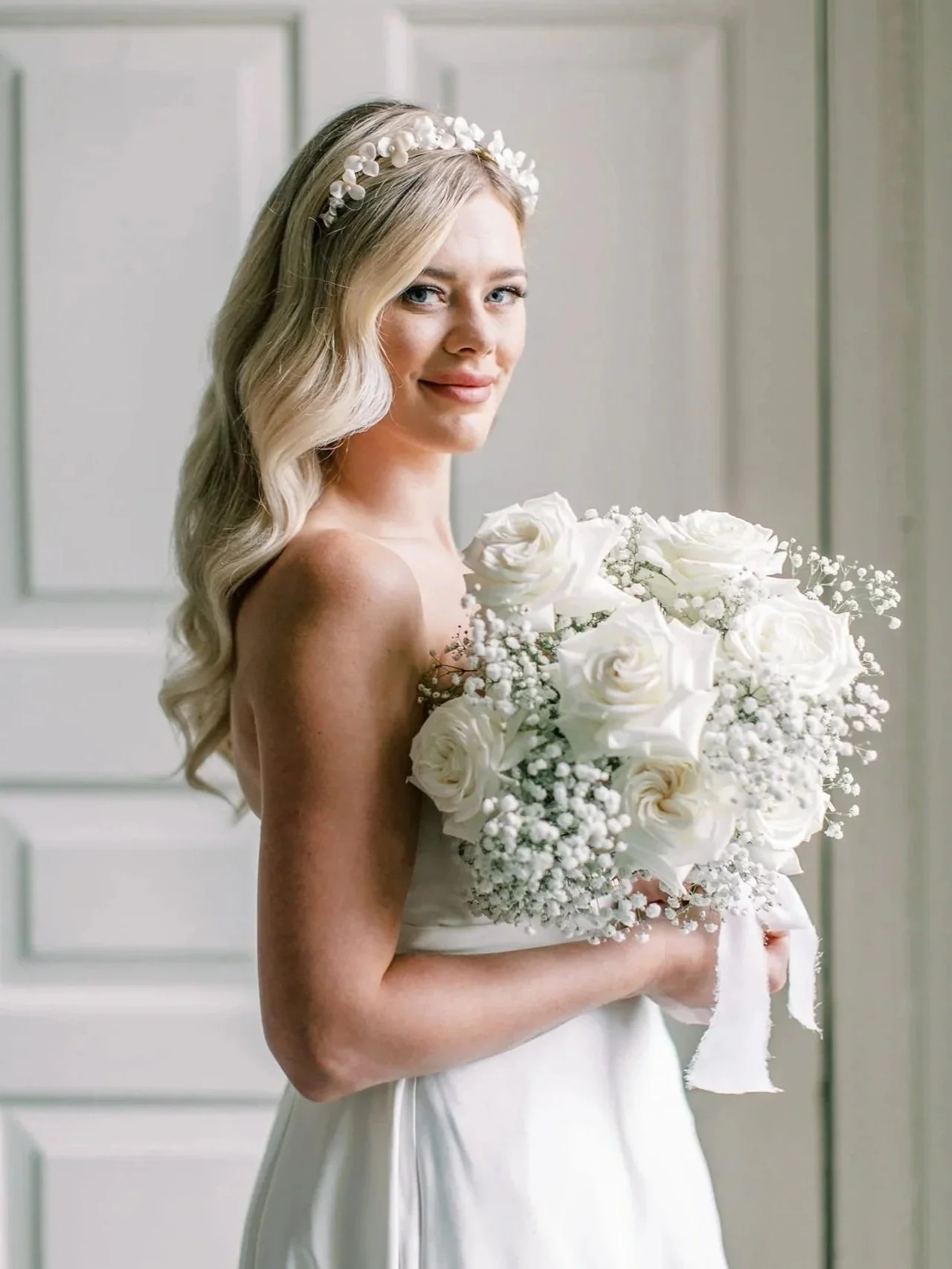 A bride with long blonde hair, wearing a floral headband, holding a bouquet of white roses and baby's breath, standing in front of a white door.