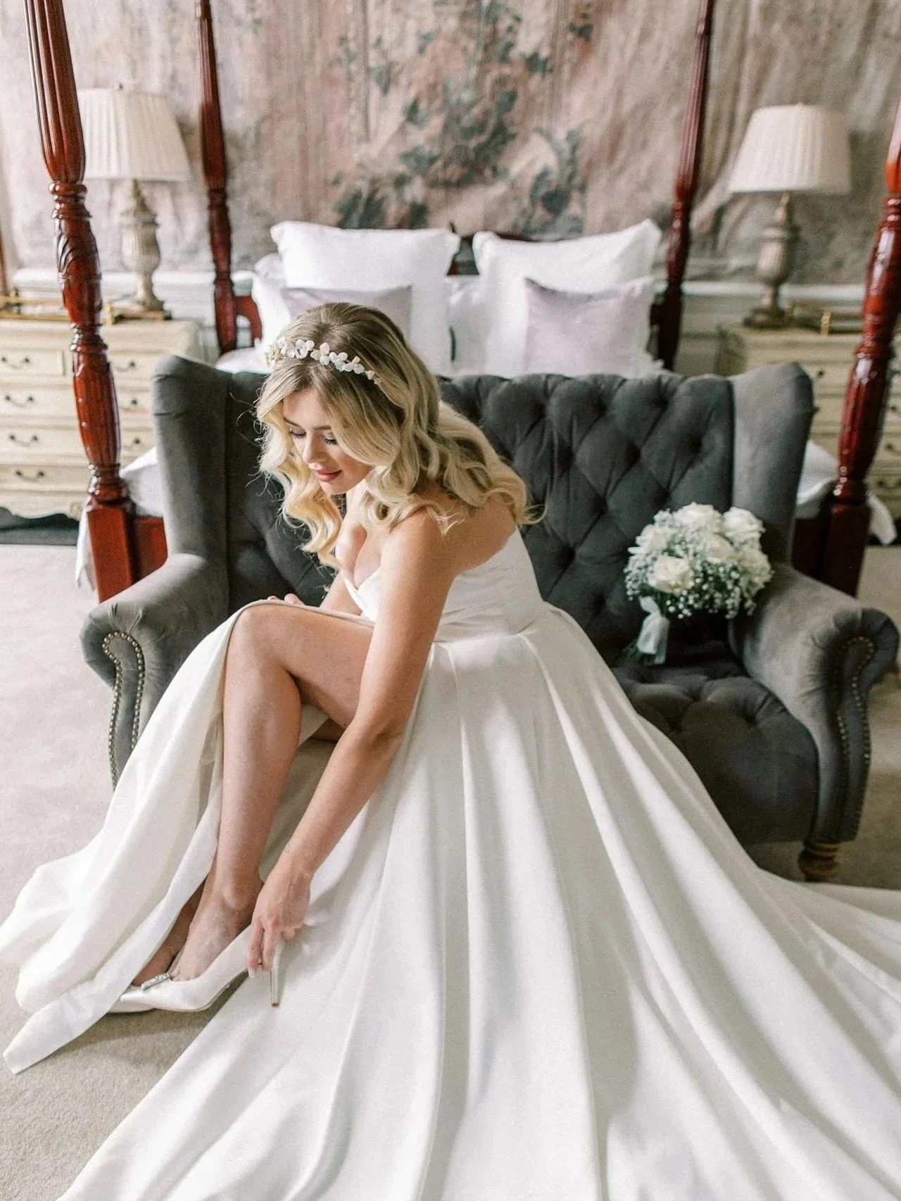 A bride in a white wedding gown sitting on a gray velvet sofa, putting on her white high heels, in a richly decorated bedroom with a four-poster bed, white bedding, and floral arrangements.