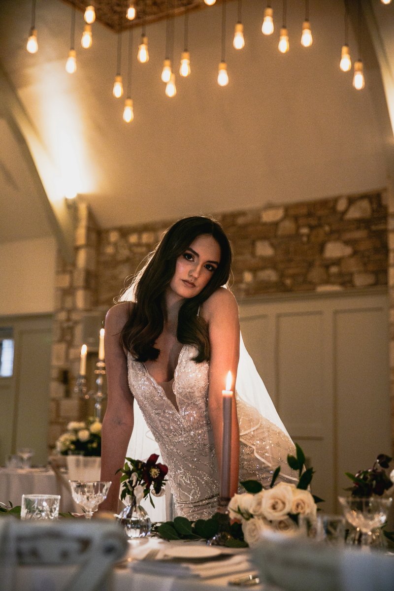 A woman in a wedding dress leaning forward at a decorated table with lit candles and floral arrangements inside a warmly lit venue with hanging bulbs and stone wall.
