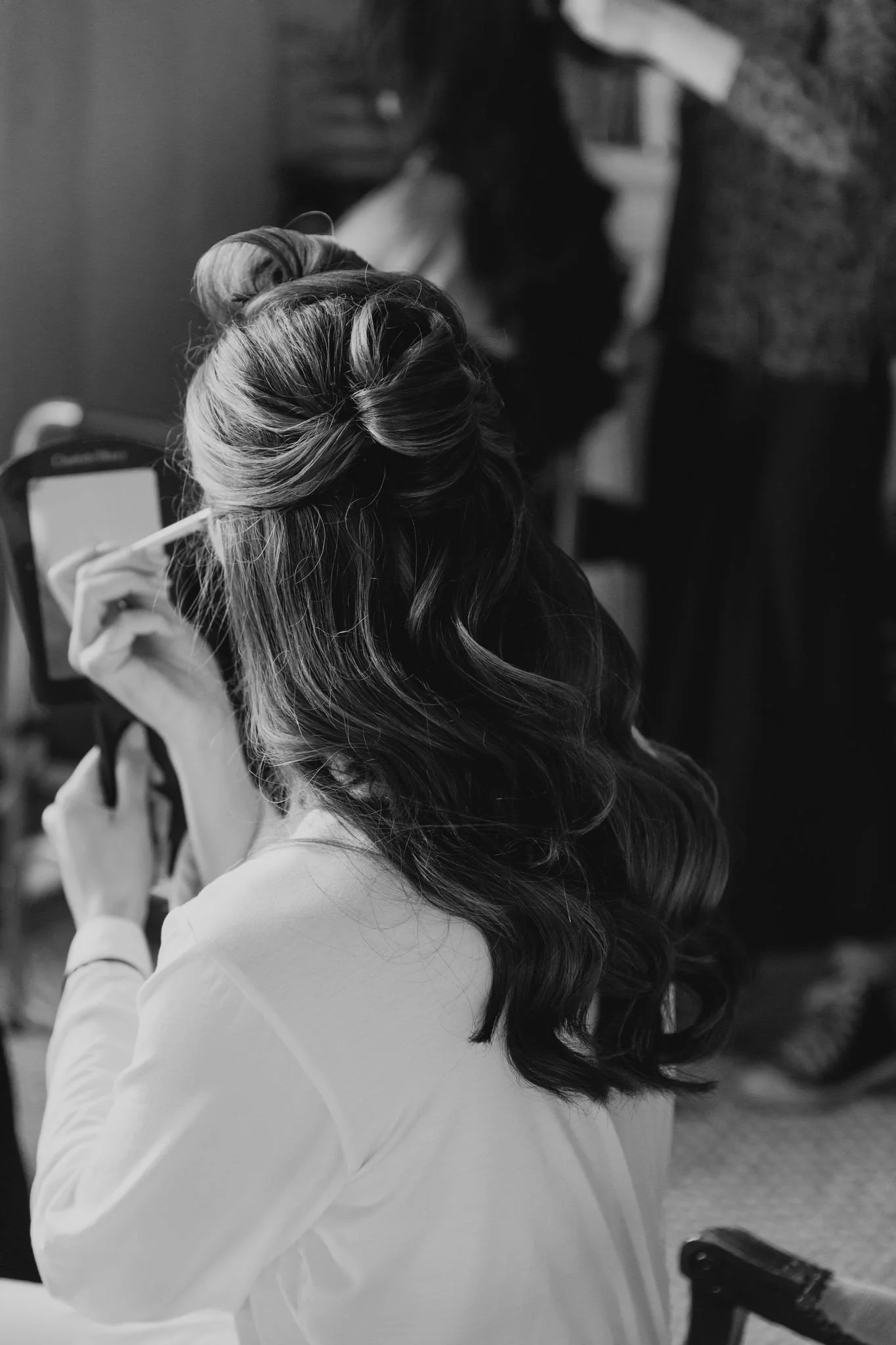 A woman with styled wavy hair in a partial updo, sitting and applying makeup with a mirror in her hand.