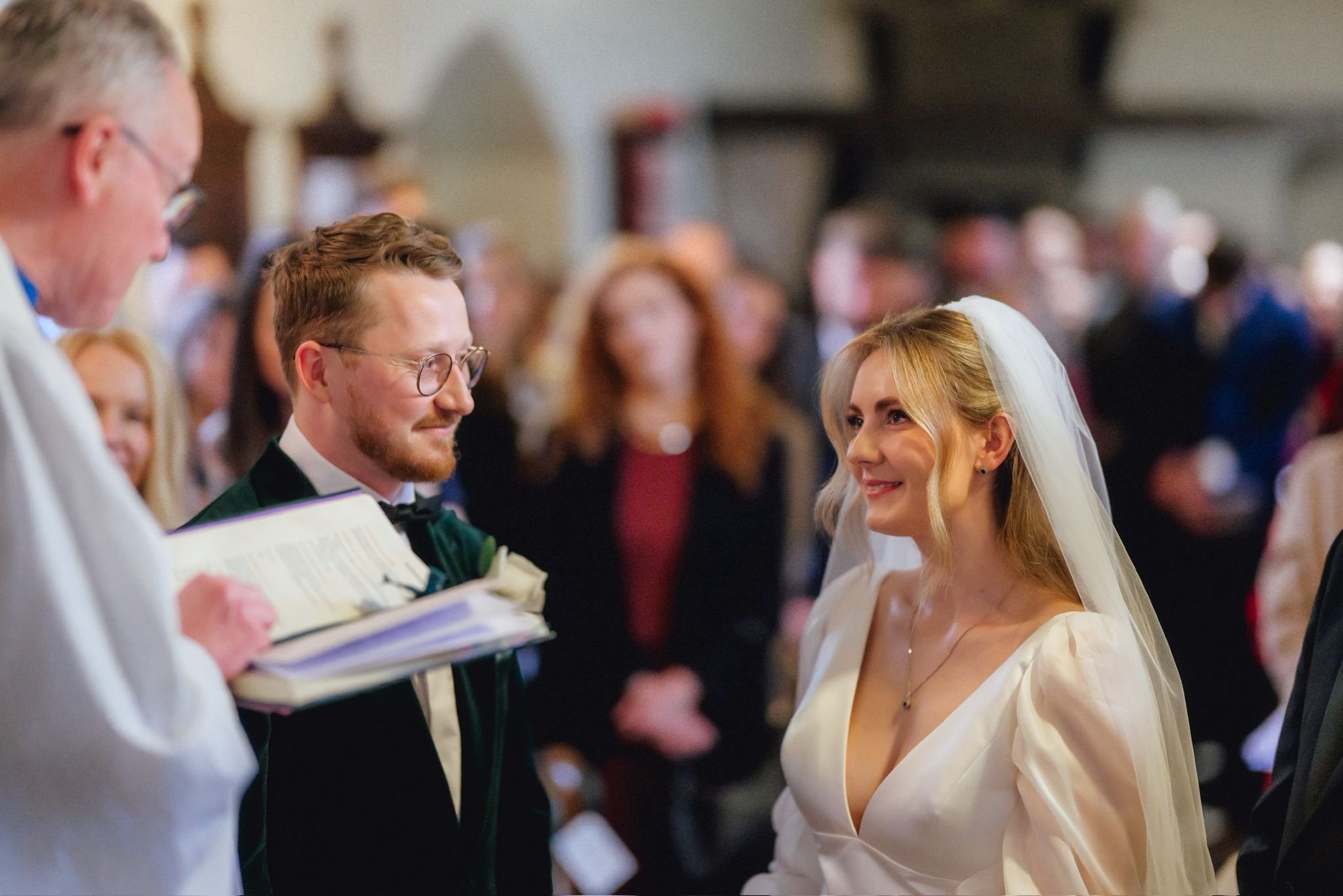 A bride and groom standing in front of a priest during a wedding ceremony, smiling at each other indoors with a blurred group of guests in the background.